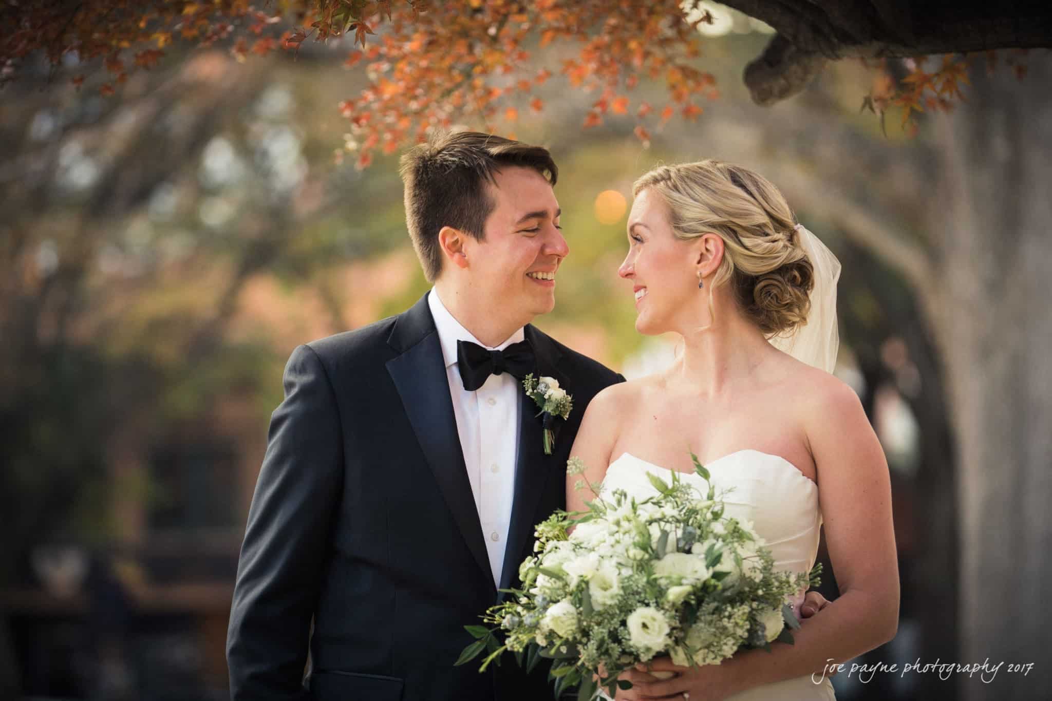 A happy couple stands outdoors on a sunny day. Both are dressed in elegant attire, with the woman holding a bouquet of flowers. They are smiling at each other under a tree with autumn-colored leaves.