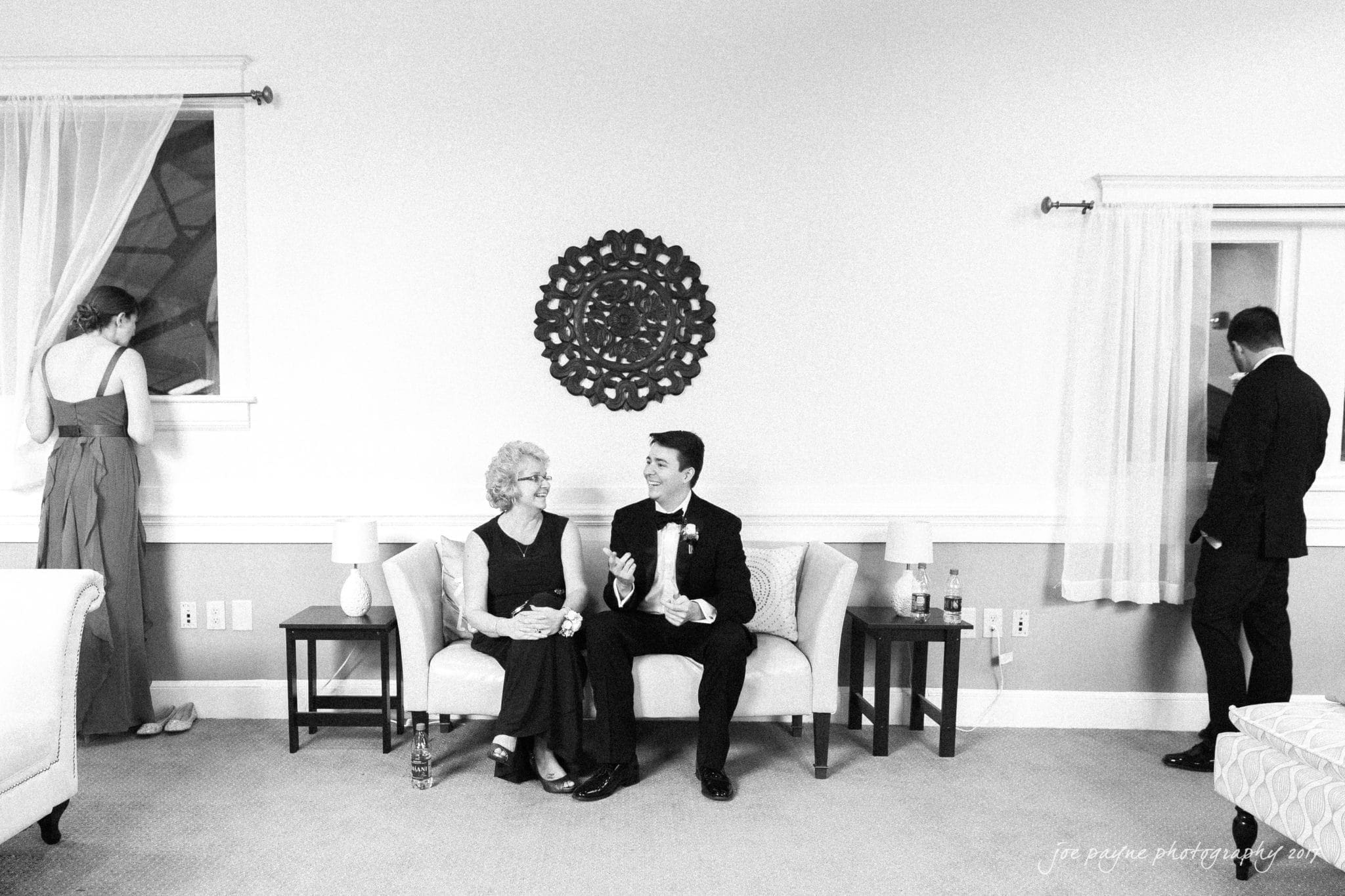 A black-and-white photo of a formal living room. An older woman sits on a couch talking with a man in a suit. Two people stand near windows, one in a dress adjusting curtains and the other looking outside. A decorative piece hangs on the wall.