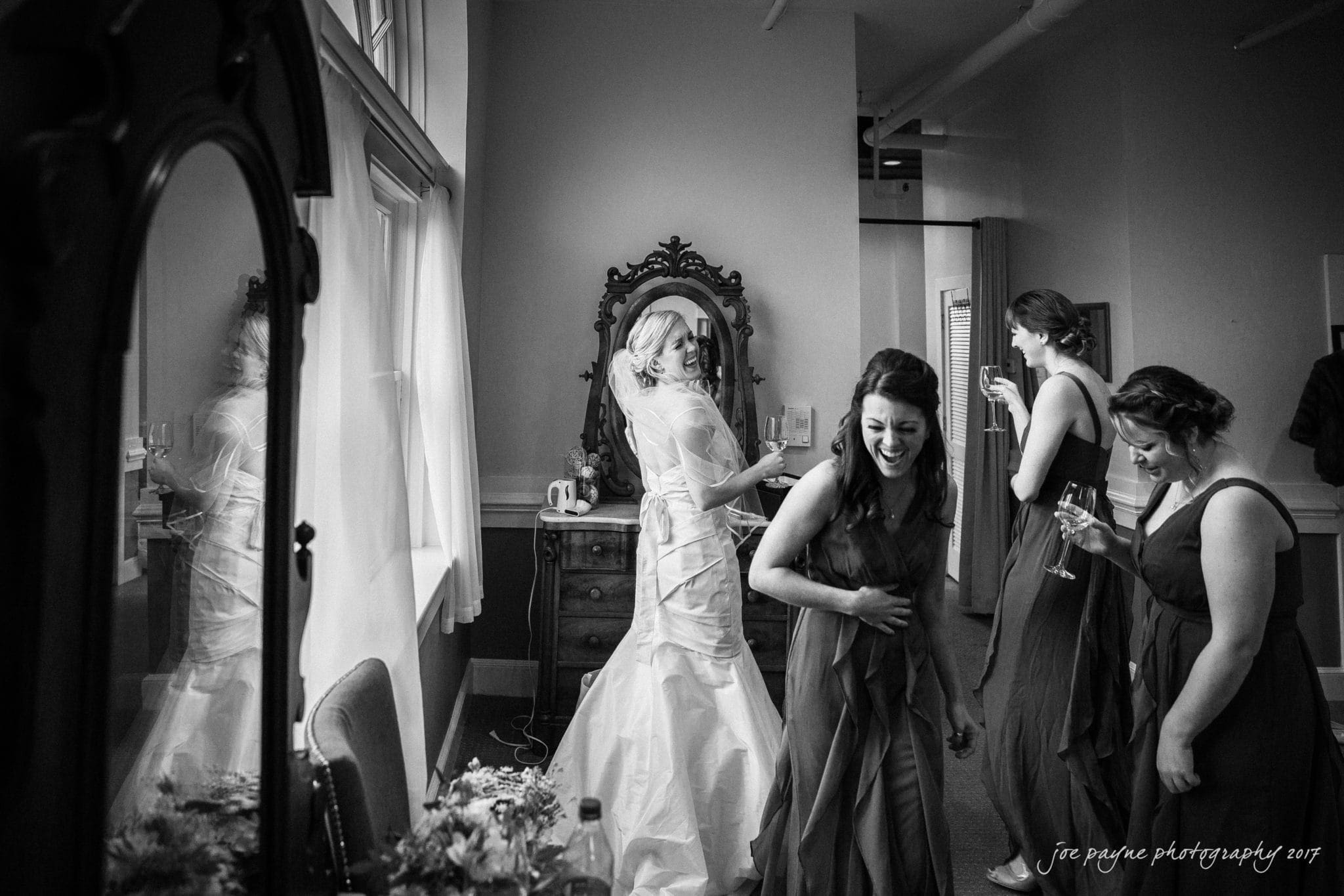 A bride in a wedding gown stands smiling in front of a mirror. Three bridesmaids in dresses are laughing and holding drinks. The room is softly lit, and the atmosphere is joyful and lively.