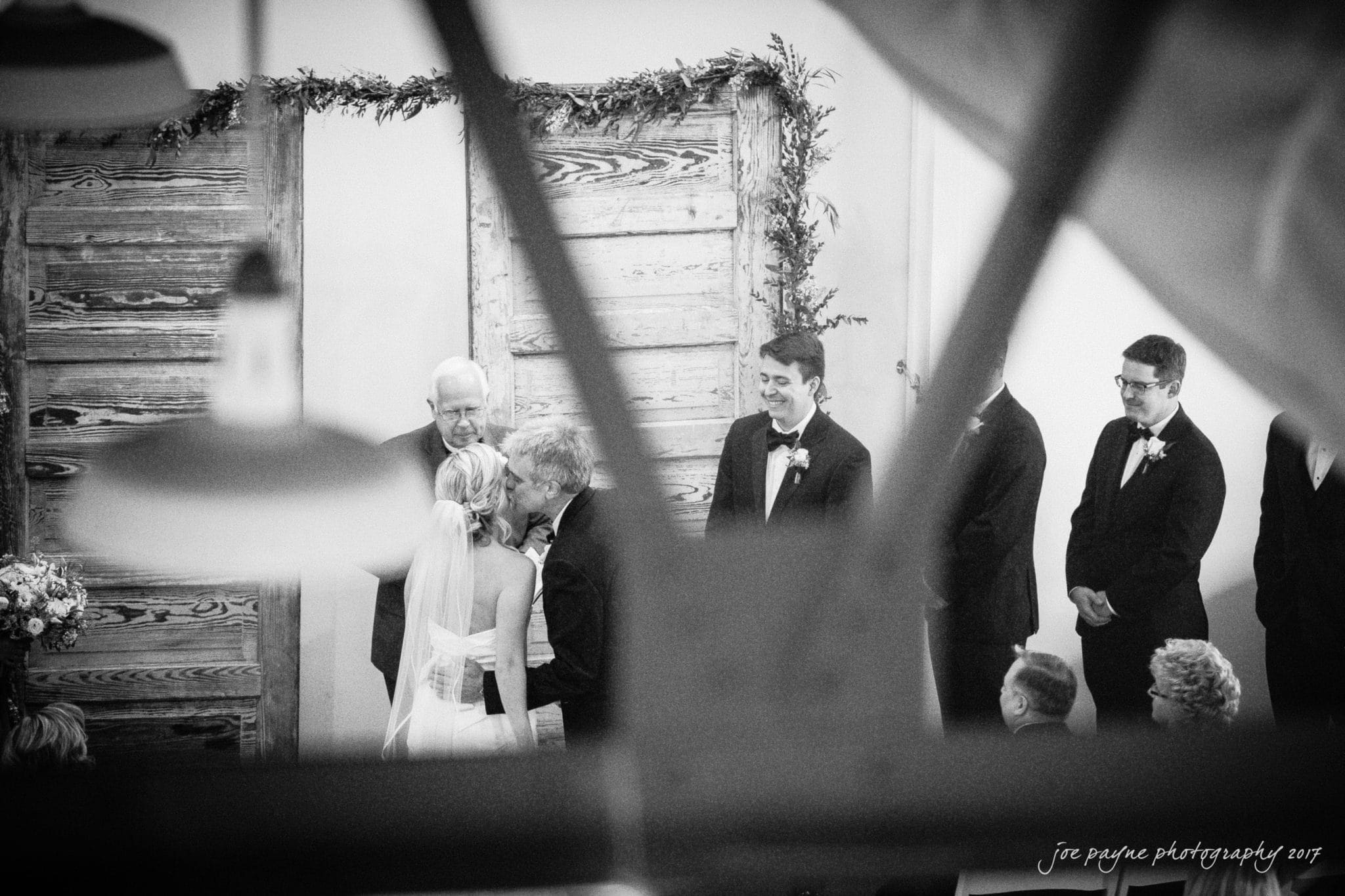 A black and white photo of a wedding ceremony. A couple stands facing each other, surrounded by three groomsmen and an officiant. The scene is set against rustic wooden doors decorated with greenery. The audience is partially visible in the foreground.