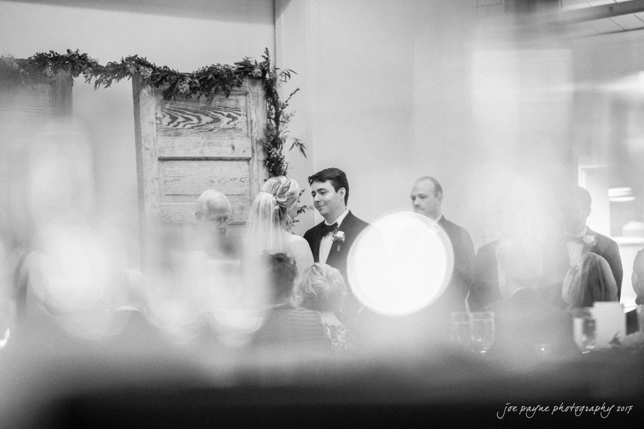 A black and white photo of a wedding ceremony. The bride and groom stand facing each other, surrounded by guests. Blurred foreground elements create a dreamy effect. The background features rustic wooden panels adorned with garlands.