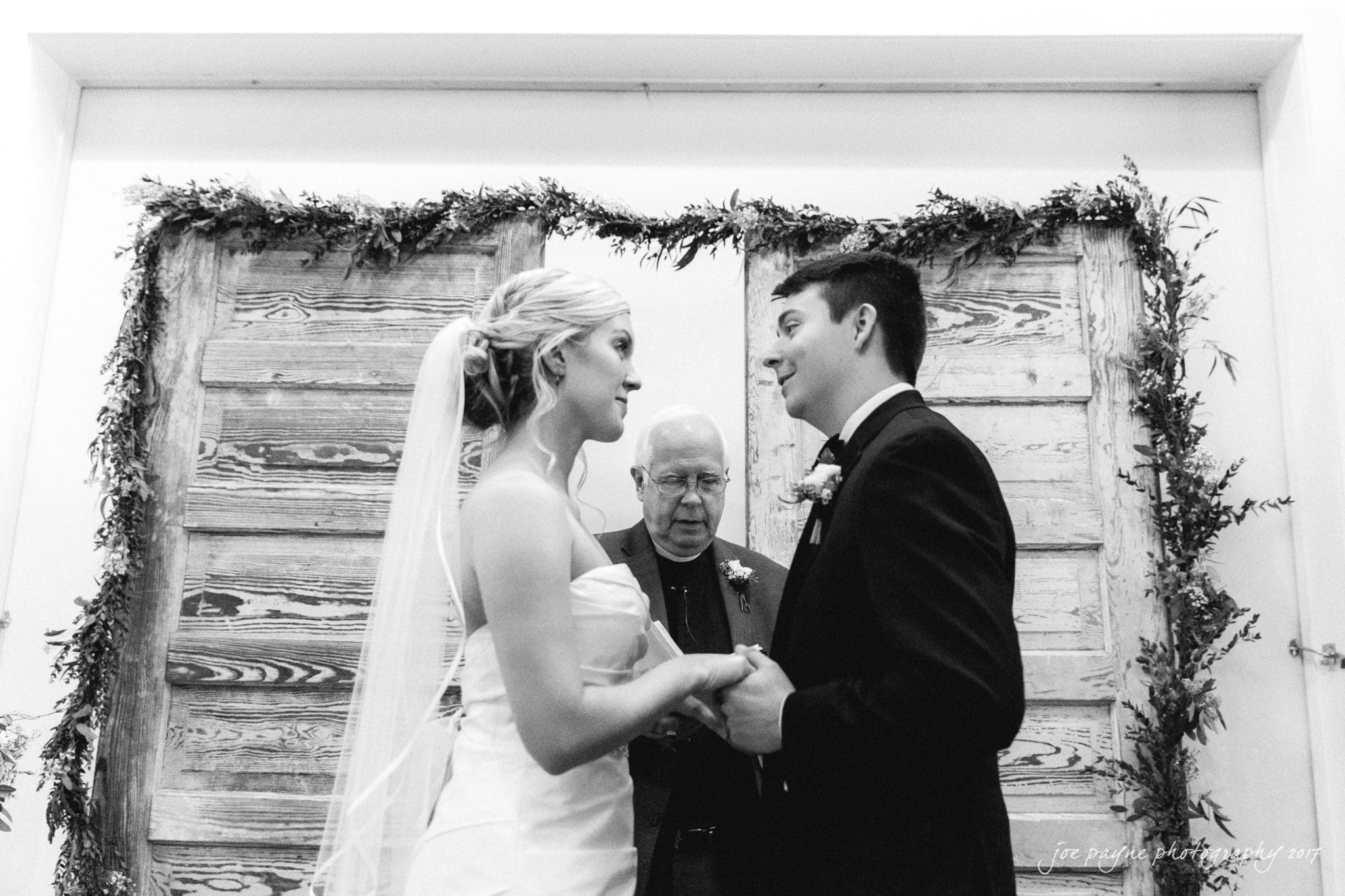 A bride and groom exchange vows during a wedding ceremony. The bride, in a strapless dress and veil, faces the groom, who is in a suit. A man stands between them, officiating, while rustic wooden doors are decorated with greenery in the background.