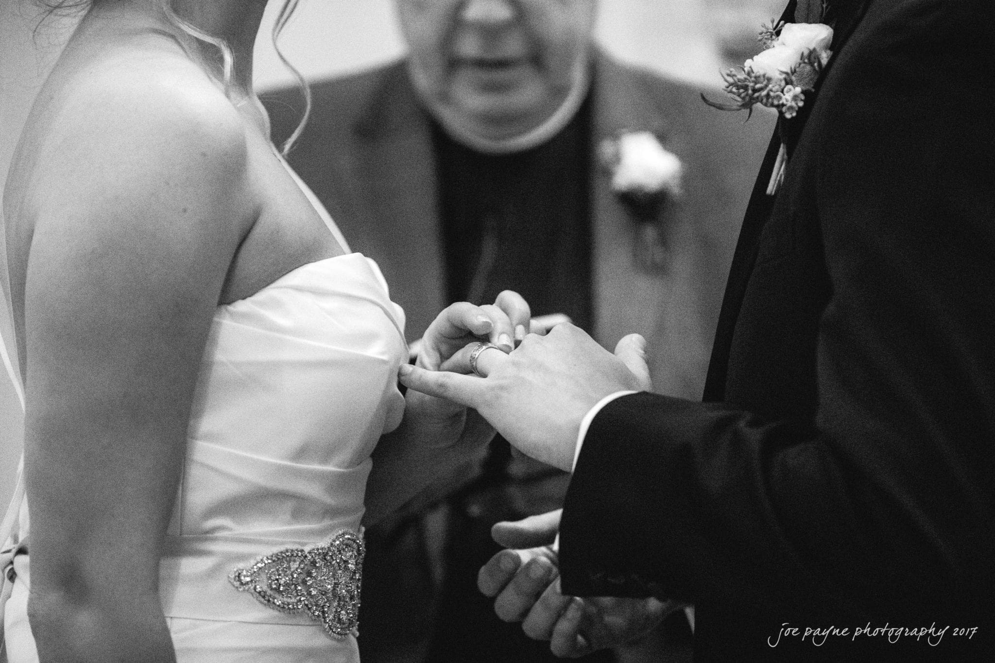 A couple exchanges rings during a wedding ceremony. The bride wears a strapless gown with an embellished waist, while the groom is in a suit. A clergyman officiates the ceremony in the background.