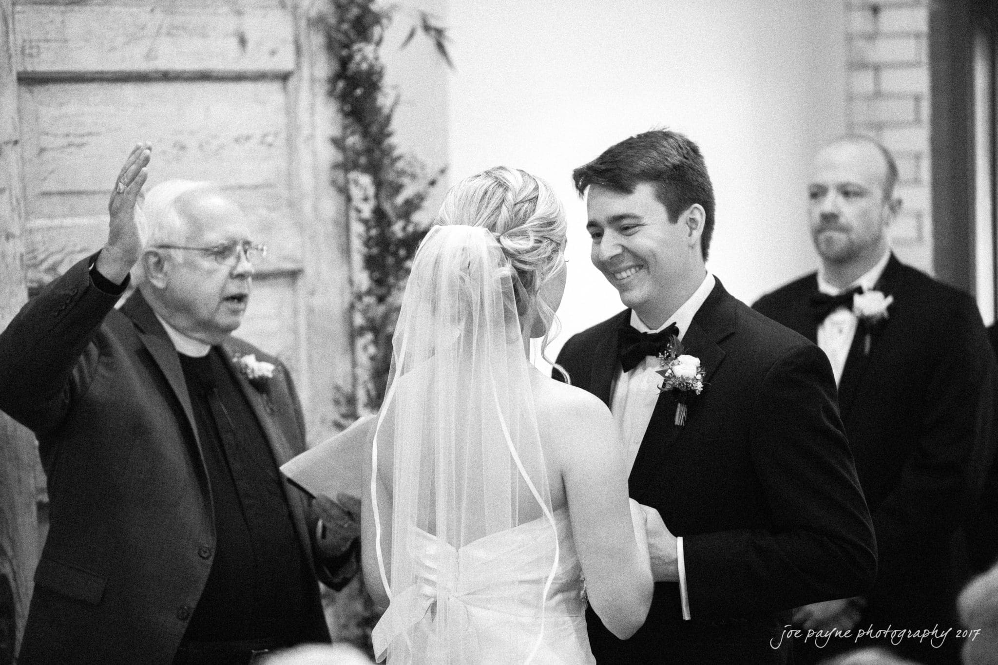 A black and white photo of a wedding ceremony. A bride and groom smile at each other, holding hands. A priest stands to their left, speaking, with another man, likely a groomsman, watching in the background.