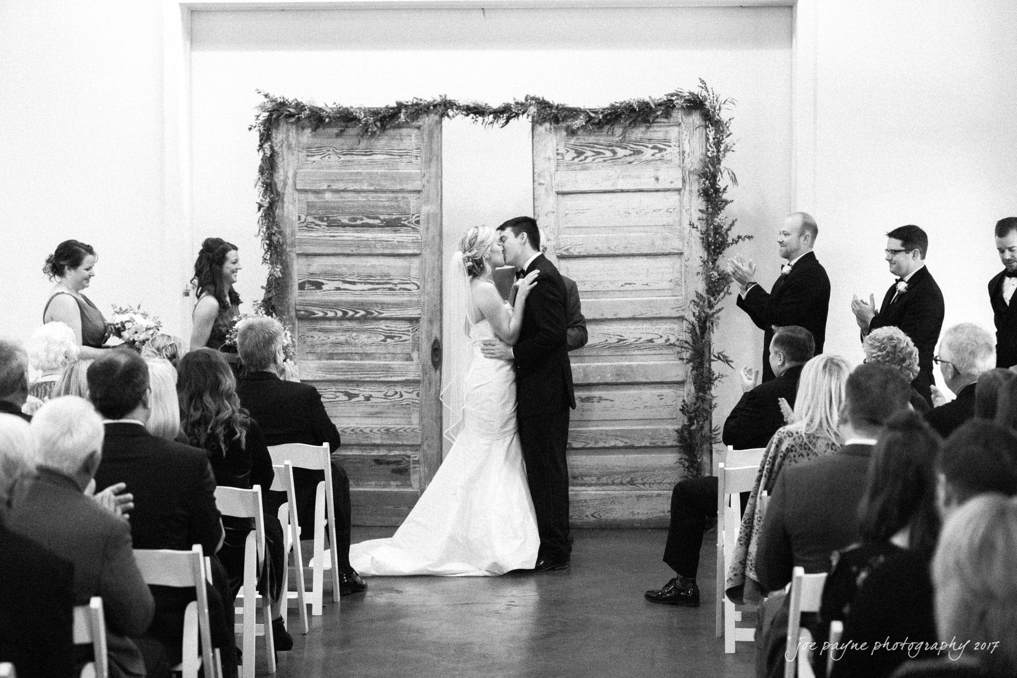 A bride and groom share their first kiss at the altar during a wedding ceremony. Guests are seated on either side, and the bridal party stands nearby. The couple is framed by rustic wooden doors adorned with greenery.