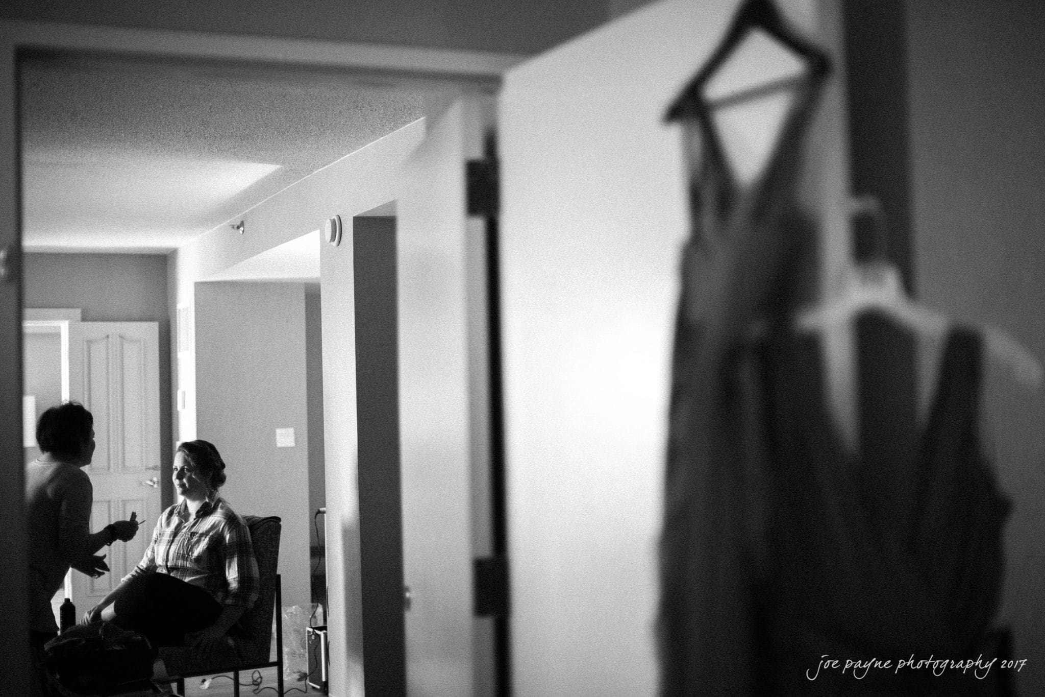 A woman sits and smiles in a room while another person applies makeup. A door partially open reveals a dress hanging on a hanger. The room is softly lit, creating a relaxed atmosphere.