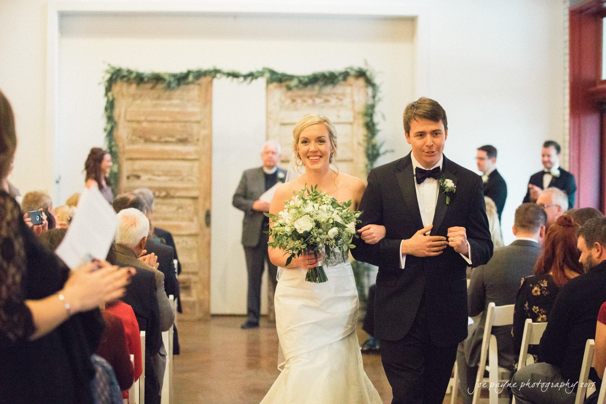 A bride and groom walk down the aisle, smiling and holding hands in a warmly lit indoor venue. The bride holds a bouquet of flowers. Guests are seated on either side, and groomsmen stand at the front. A rustic set of double doors is visible behind them.