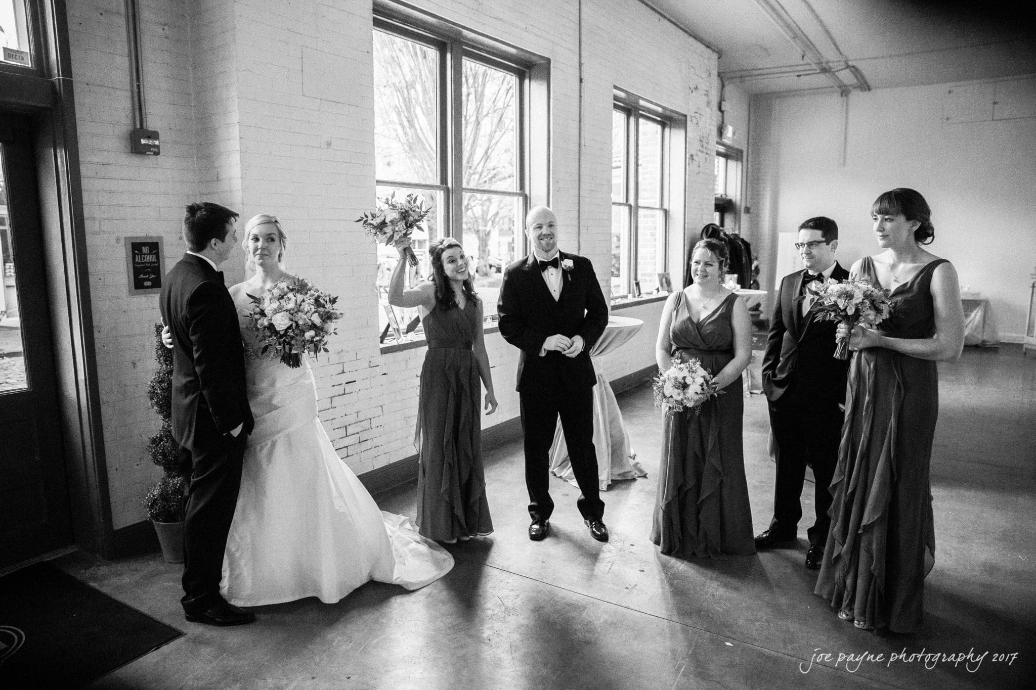 A bride and groom stand with their wedding party inside a room with large windows. The bride holds a bouquet, and another woman raises her bouquet in celebration. Everyone is dressed formally in gowns and suits.