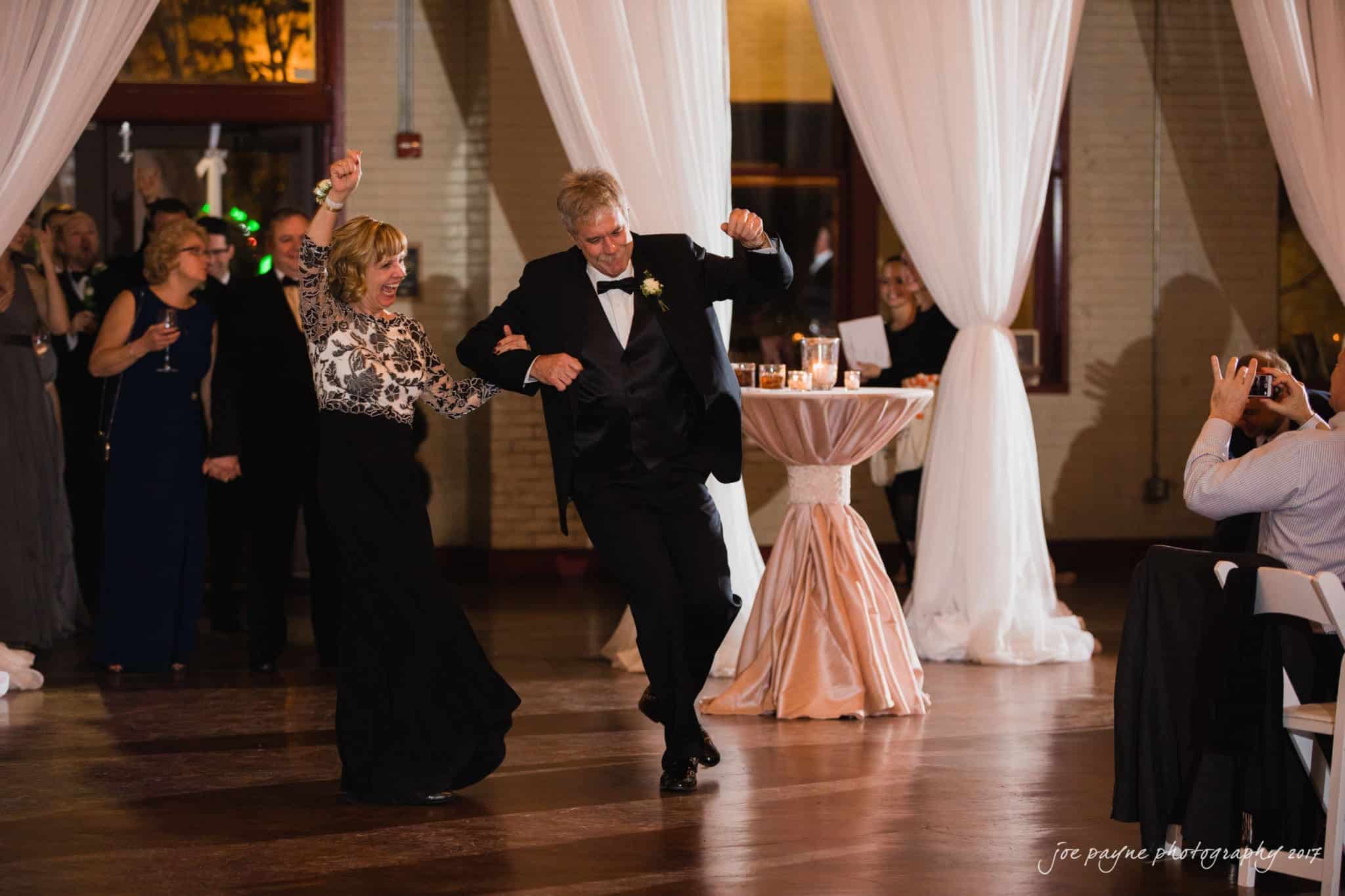 A couple in formal attire dances joyfully at a lively event. They are surrounded by elegantly dressed guests, gathered in a warmly lit venue with draped curtains and round tables.