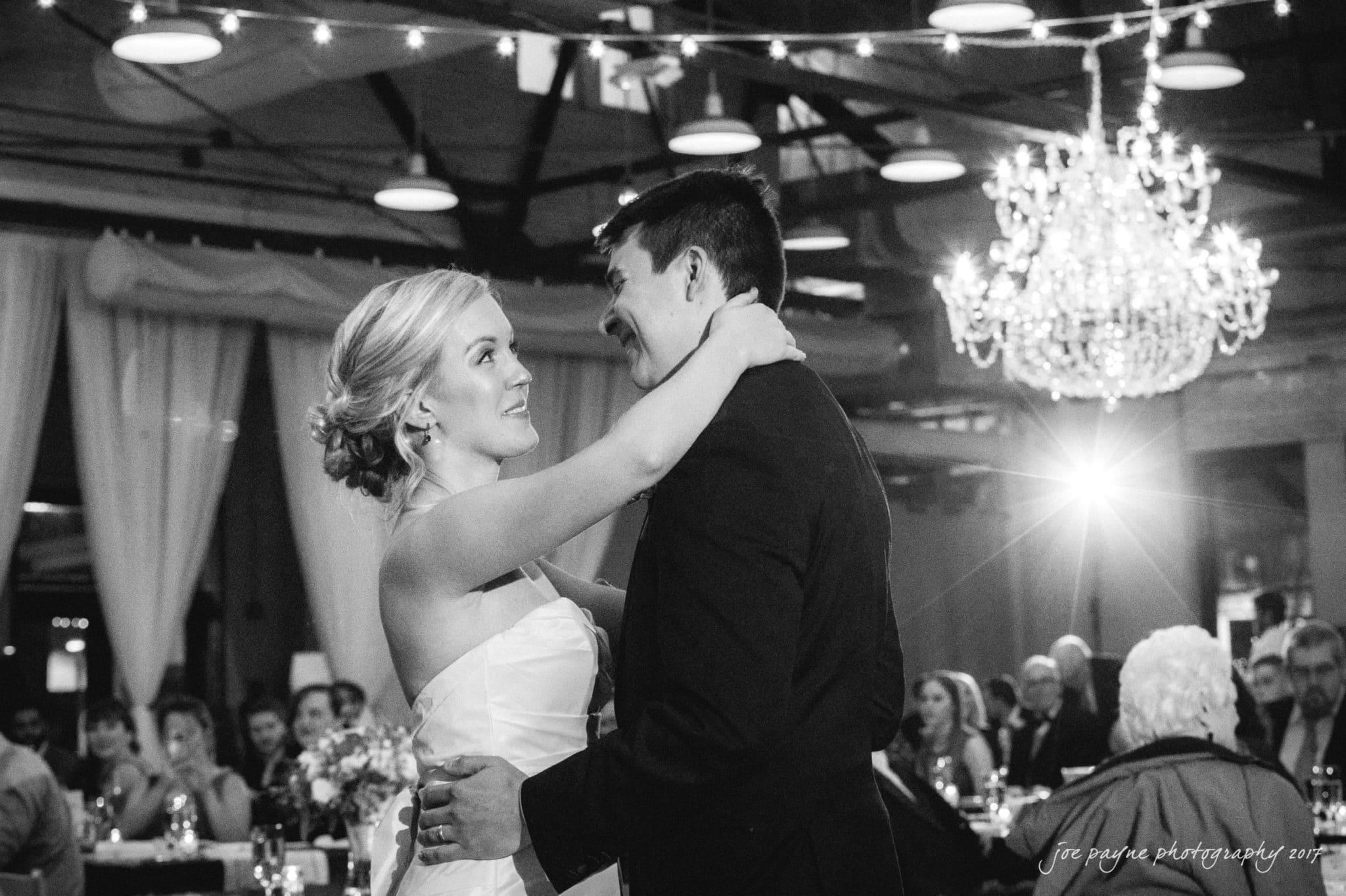 A bride and groom share their first dance under a chandelier. They are surrounded by seated guests in a warmly lit reception venue.