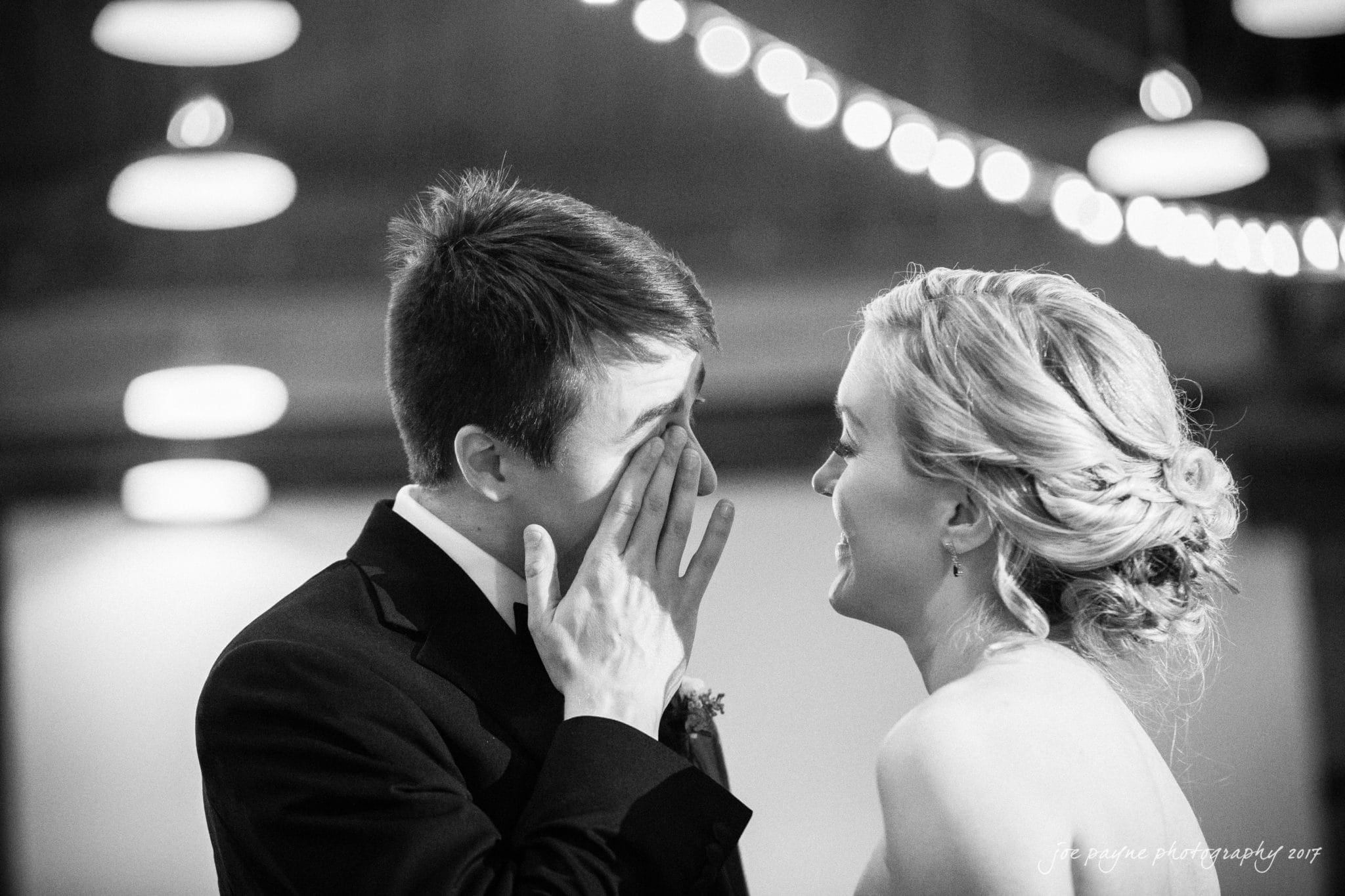 A black and white photo of a wedding couple sharing an emotional moment. The groom covers his face with his hands, appearing moved, while the bride smiles tenderly at him. Soft, out-of-focus lights twinkle in the background.