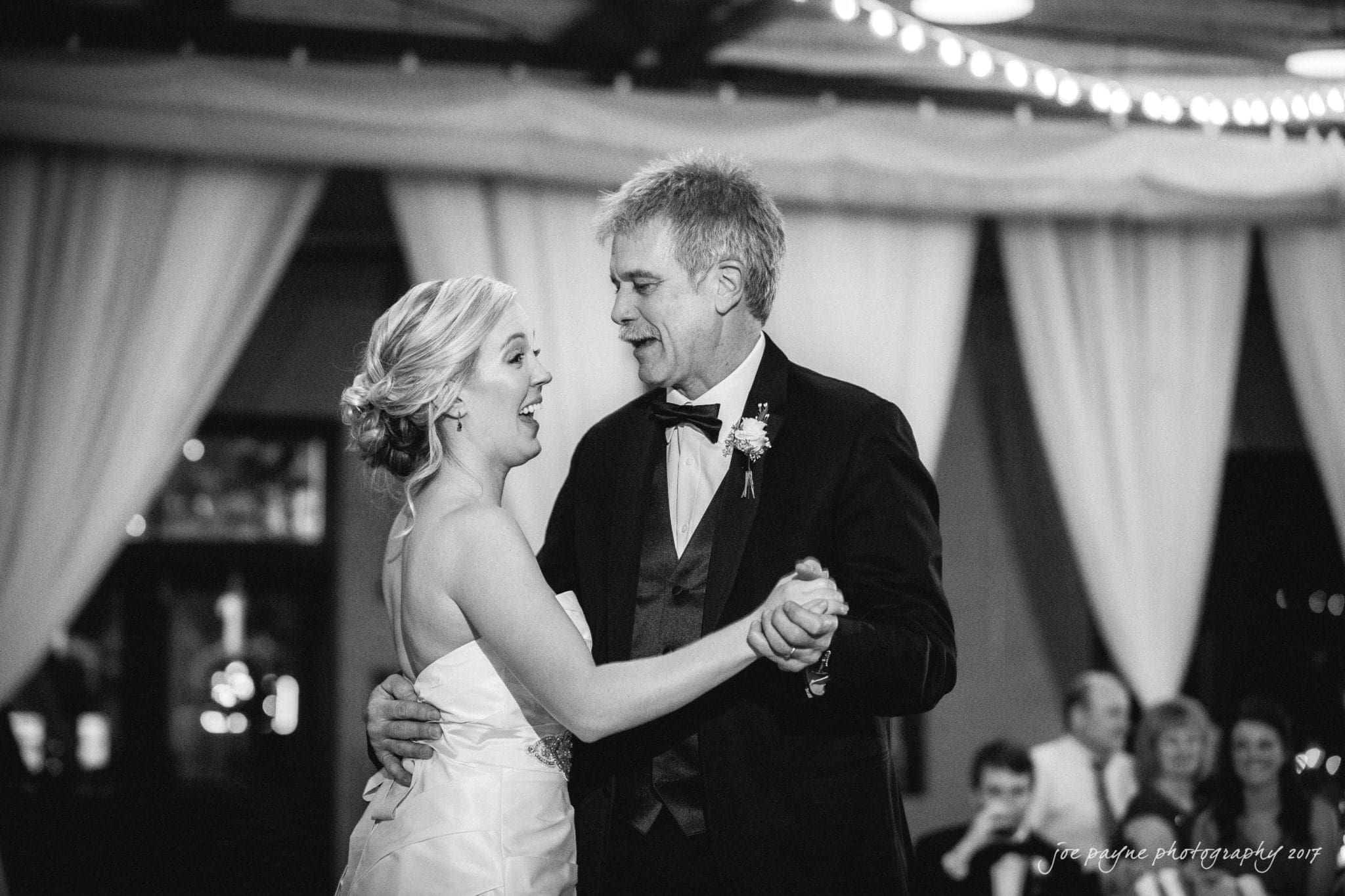 A black and white photo of a man in a suit and a woman in a white gown dancing together in a warmly lit indoor setting. They appear joyful, with people seated and watching in the background.