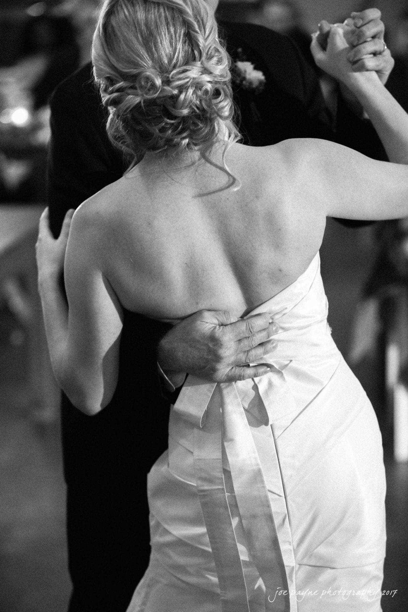 A couple dances closely, with the woman wearing a strapless white dress and her hair in an elegant updo. The man, in a dark suit, embraces her while leading the dance. The setting appears to be a warmly lit indoor venue.