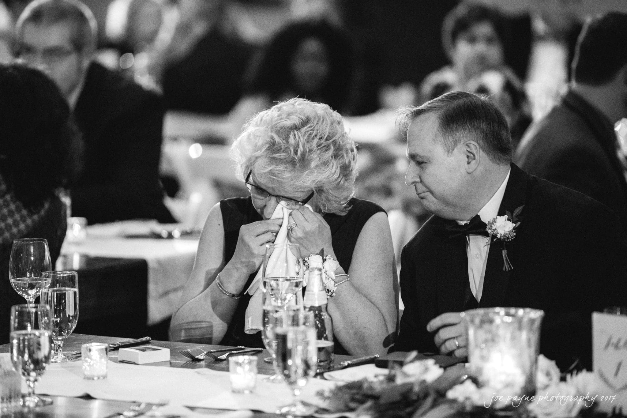 A woman wipes her tears with a tissue while a man sits beside her, looking on. They are at a table set with glasses and candles, surrounded by other people in a dimly lit setting, possibly a formal event or celebration.