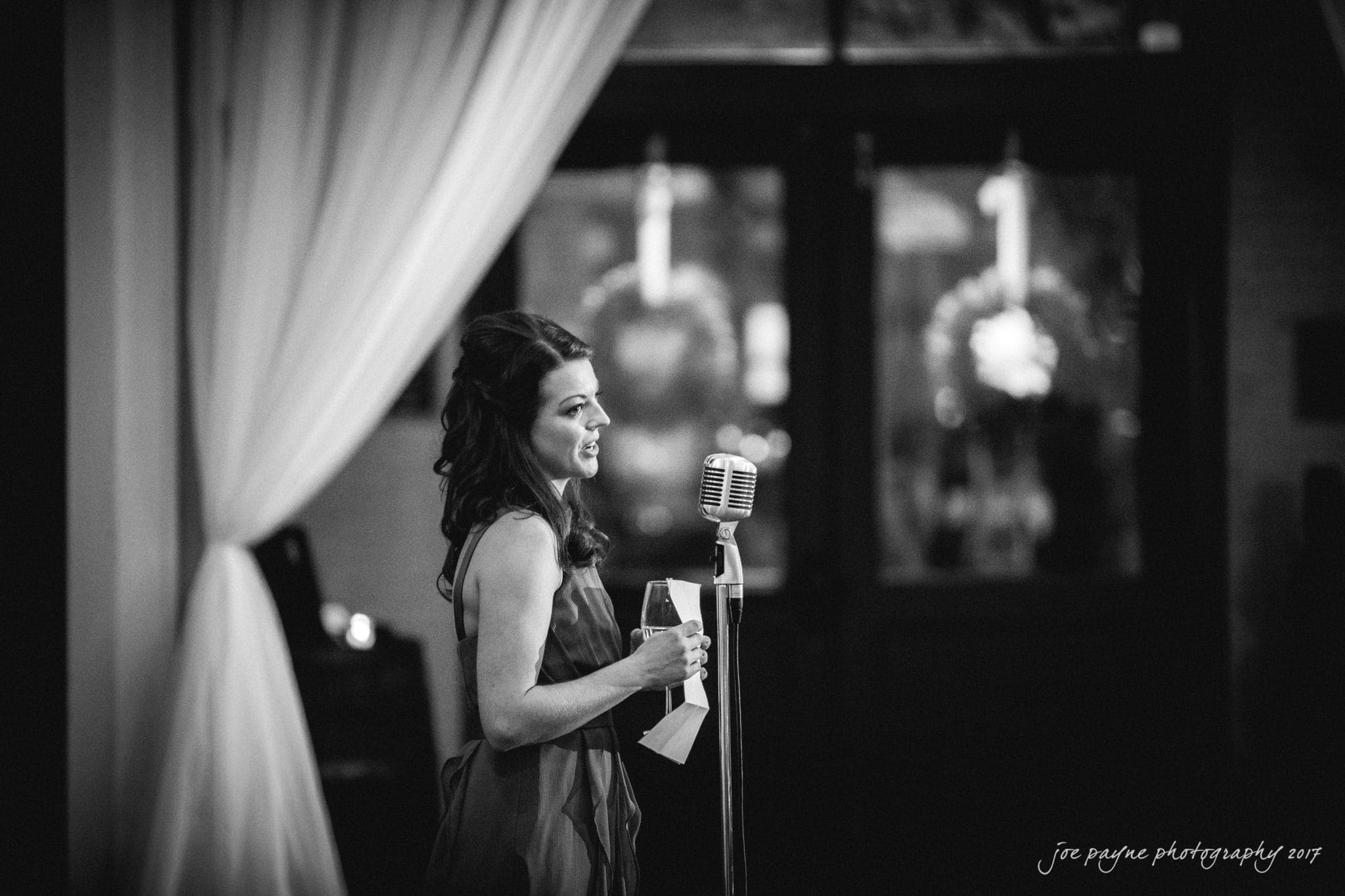 A woman stands with a vintage microphone in front of white drapes, holding a piece of paper and a drink. The black and white image captures her mid-speech, creating an elegant and nostalgic atmosphere.