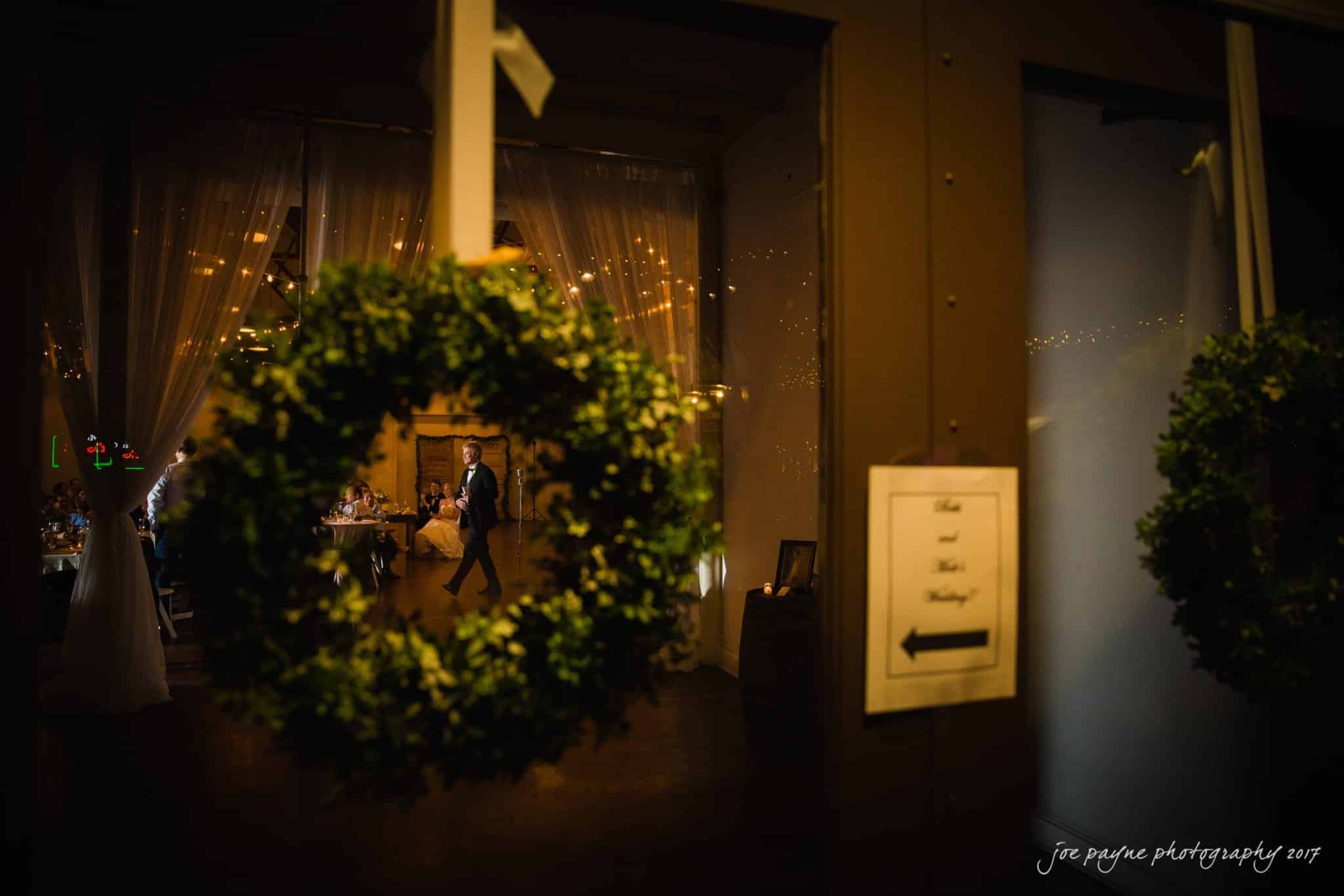 A wedding reception with dim lighting, featuring a couple dancing in the background. Green wreaths hang on a glass door with a sign pointing to the wedding venue. Soft lights and draped curtains create a warm ambience.