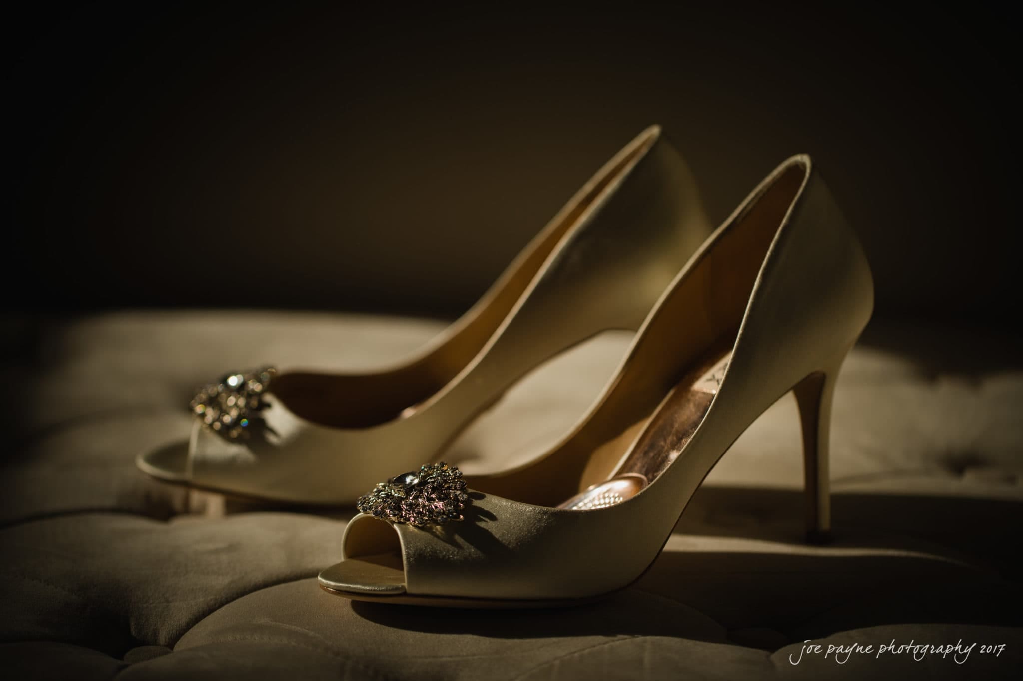 Elegant high-heeled shoes with jeweled embellishments on the open toes, displayed on a tufted surface under soft lighting.