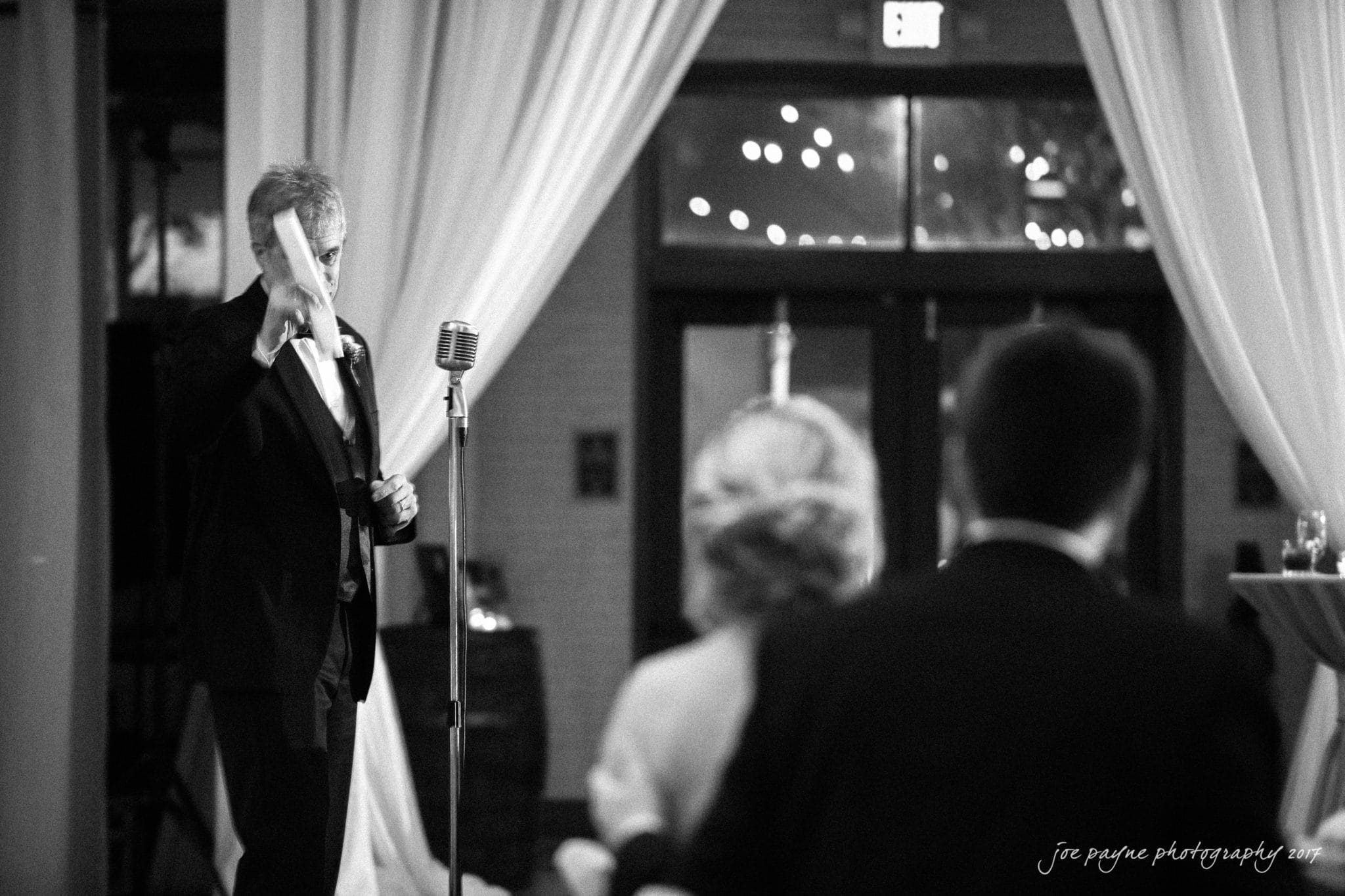 A black and white photo of a man in a suit giving a speech with a microphone and raised hand in a dimly lit room. Curtains and lights are visible in the background. An audience is partially visible in the foreground.
