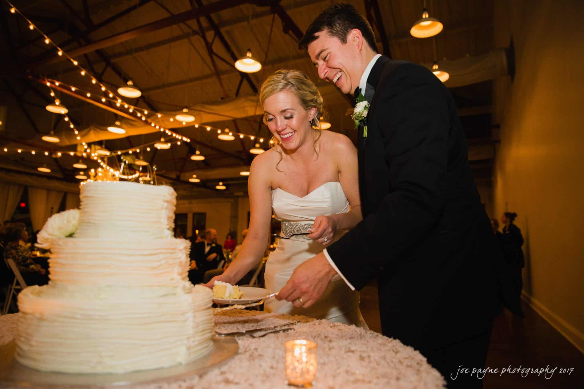 A bride and groom smile as they cut a multi-tiered white wedding cake. The bride wears a strapless gown, and the groom is in a suit with a boutonniere. The setting is a warmly lit reception hall with guests seated in the background.