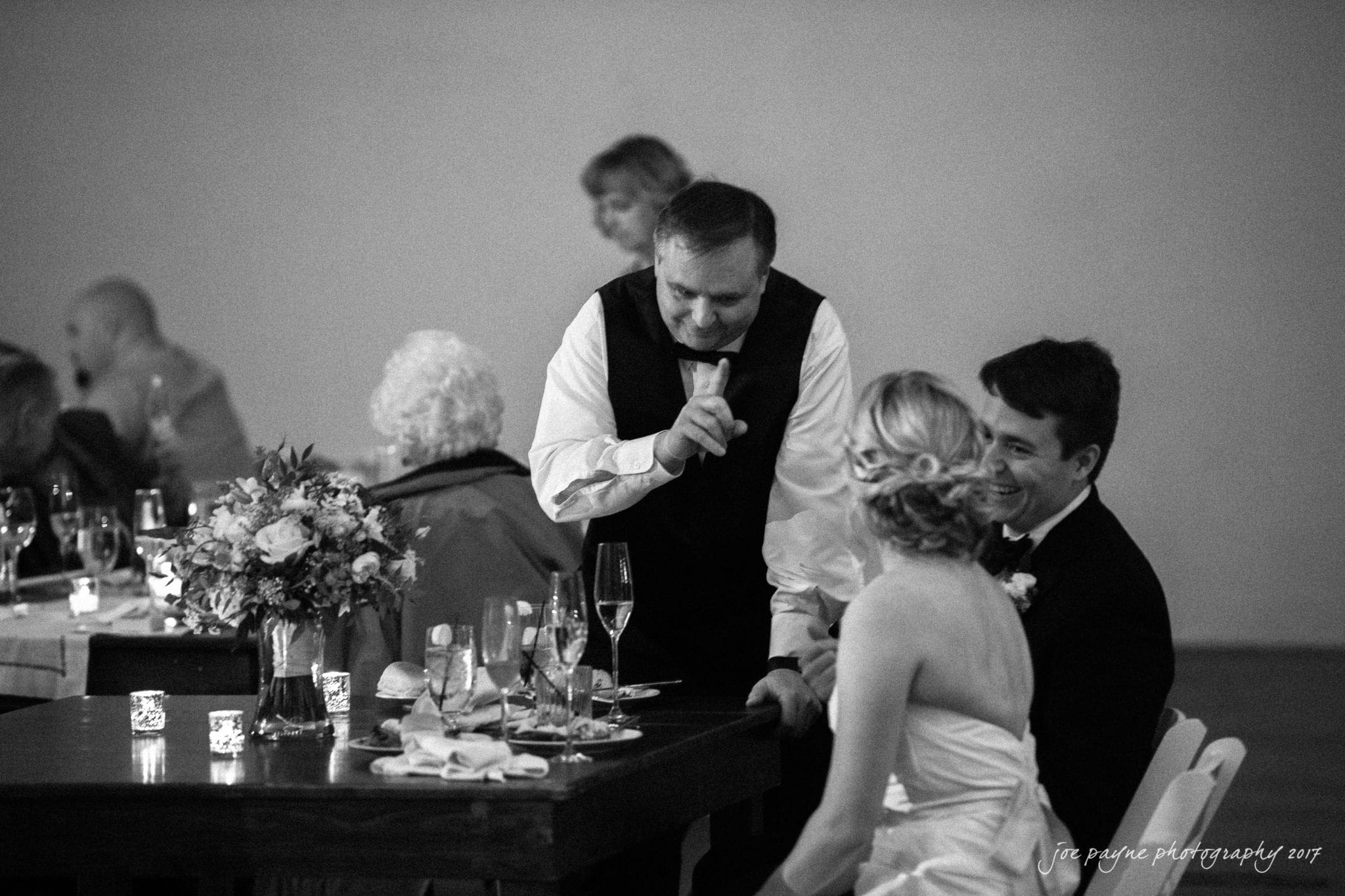 A man in a vest gestures with a finger to his lips, signaling quiet, as he stands near a seated couple in formal attire at a dining table. Other guests are visible in the background, and flowers decorate the table.