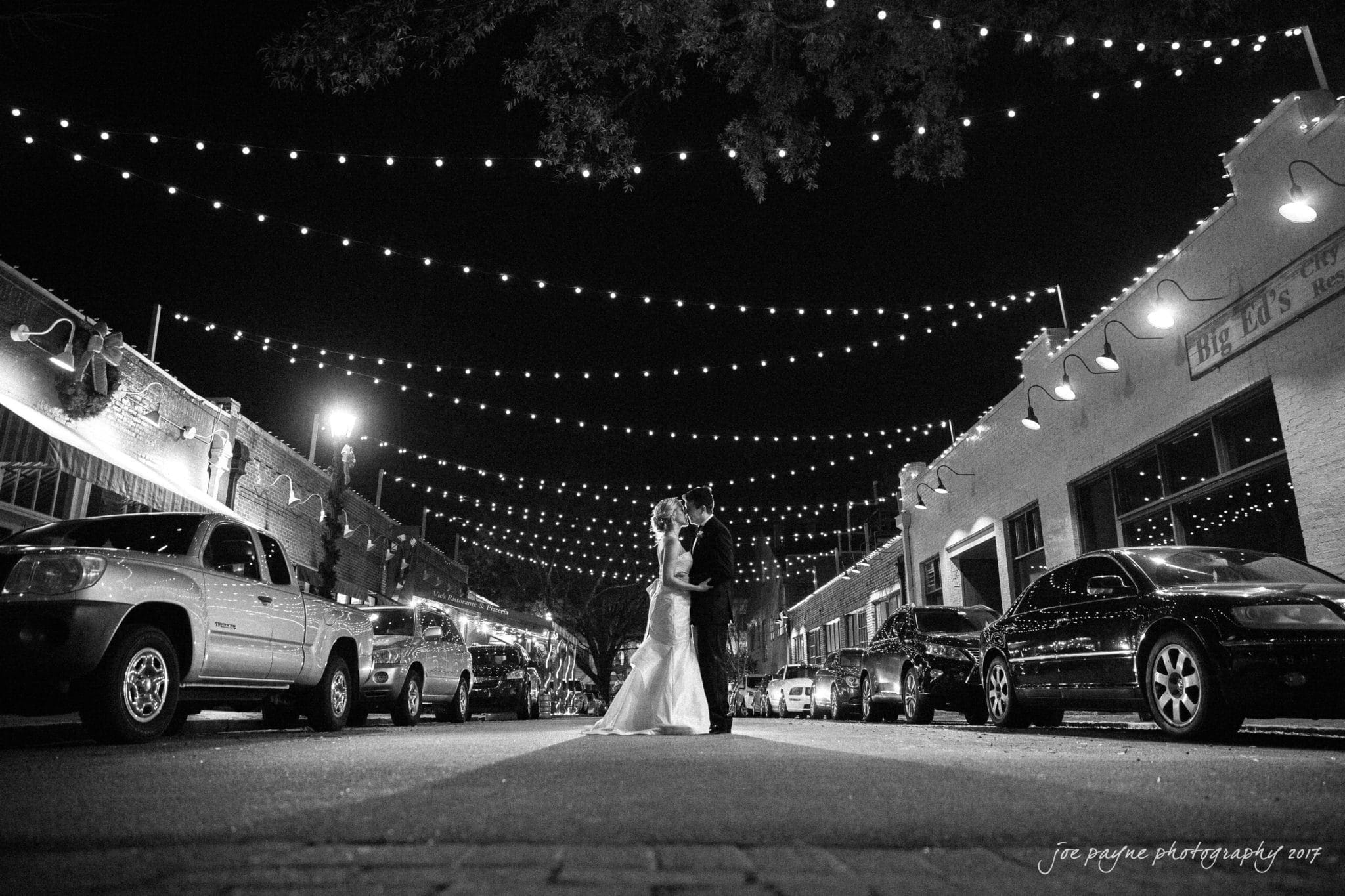 A black-and-white photo of a couple in formal attire embracing in the middle of a street at night. String lights are overhead, and cars line both sides of the street. The scene is romantic and atmospheric.