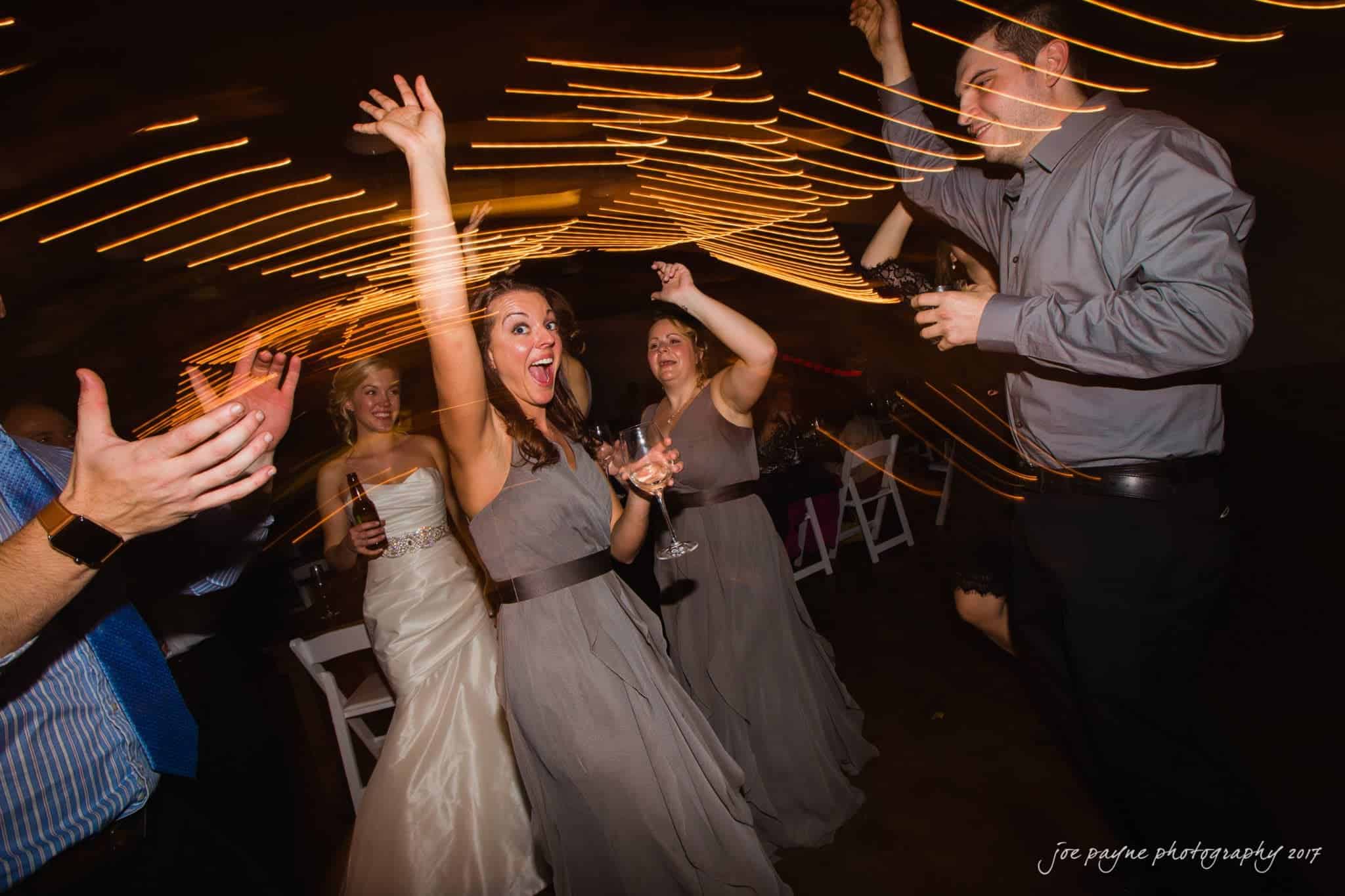 A group of people dancing energetically at a wedding reception. The woman in the center raises her arms joyfully, surrounded by other guests in formal attire. String lights create a dynamic effect around them.