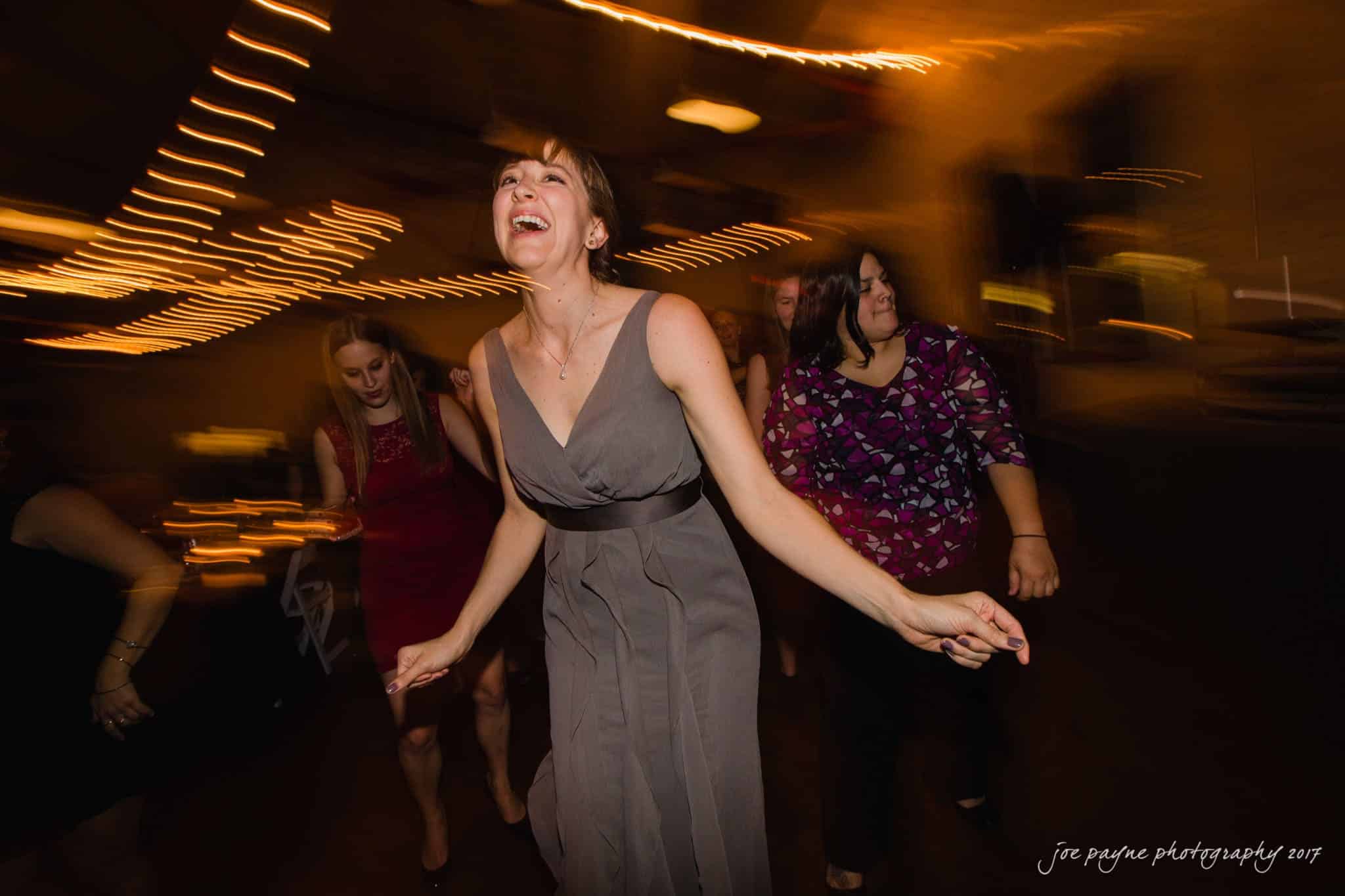 A group of people dance energetically at a party. A woman in a gray dress is prominently visible, smiling with joy. String lights create a warm, festive atmosphere. Motion blur adds a lively, dynamic element to the scene.