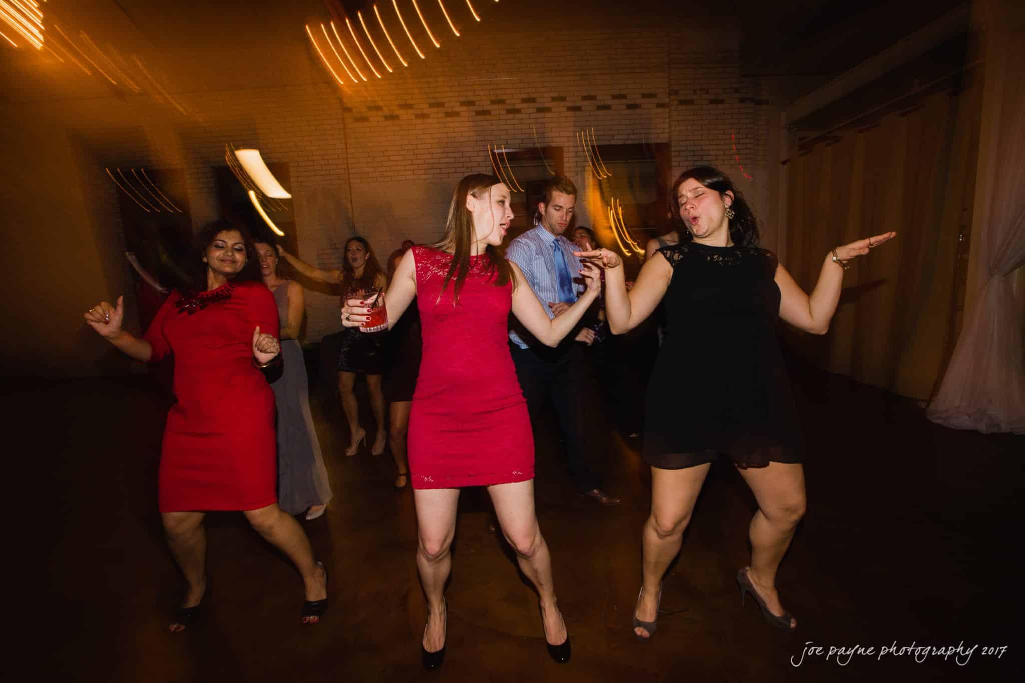 A group of people dancing energetically at a dimly lit indoor event. Two women in red and black dresses are in the foreground, and others are enjoying the moment in the background. Warm lights create a lively atmosphere.
