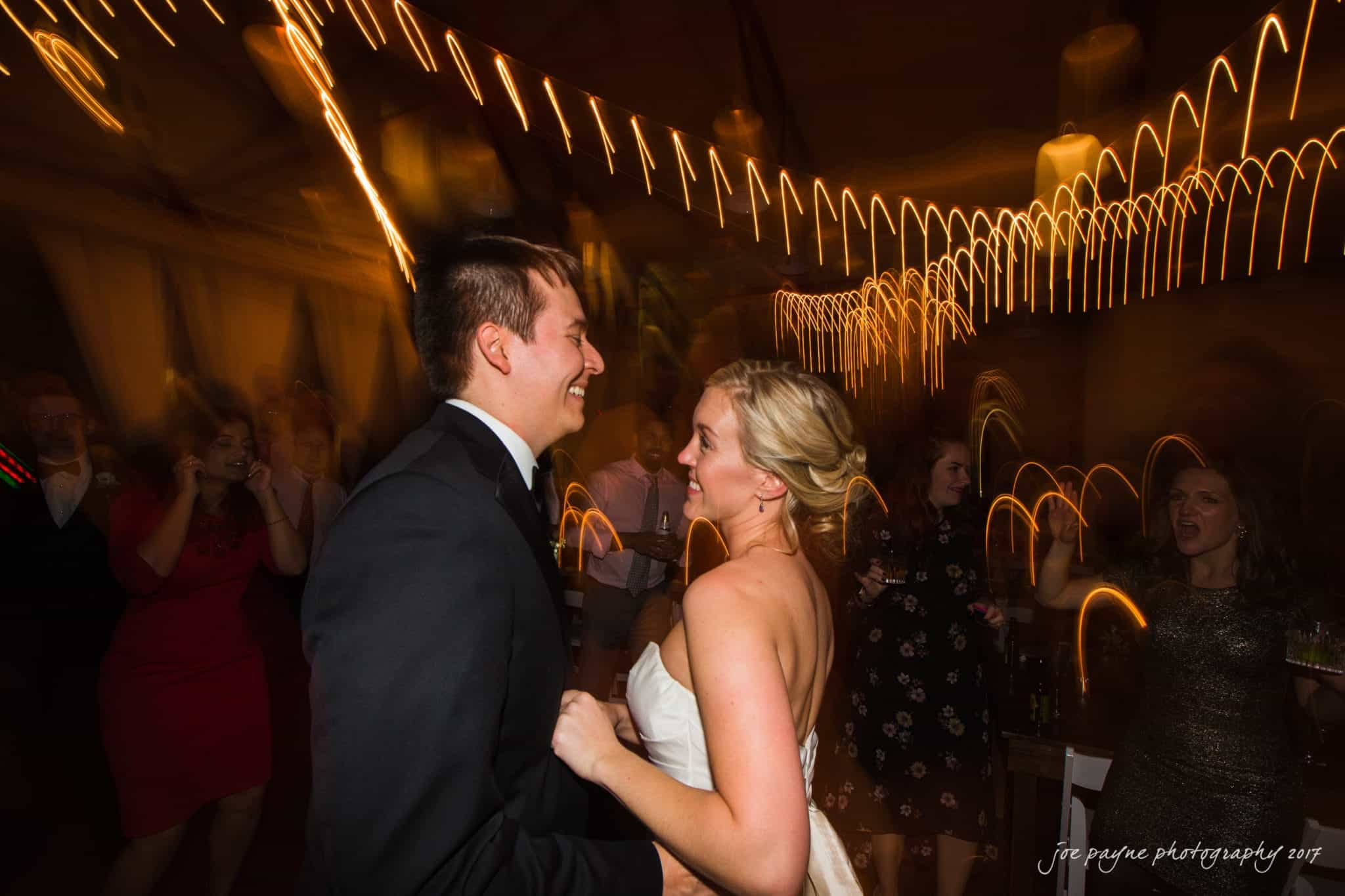 A couple dances joyfully at a wedding reception. The woman wears a white gown, and the man is in a dark suit. The dimly lit room features guests in the background with blurred lights creating an energetic atmosphere.