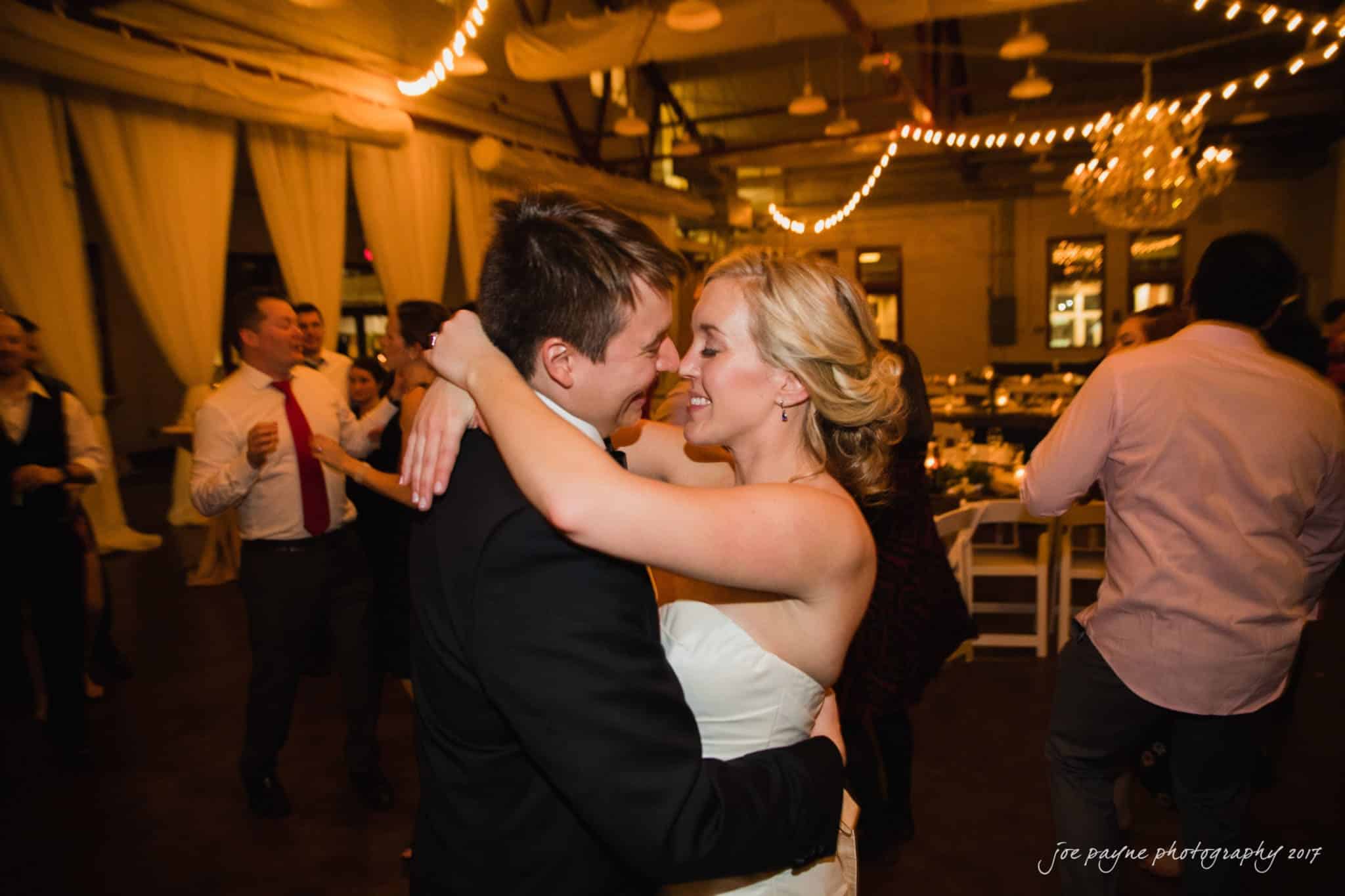 A couple dances closely in a warmly lit indoor venue, surrounded by guests. The woman wears a white dress, and the man is in a dark suit. String lights and elegant drapery add to the festive atmosphere.