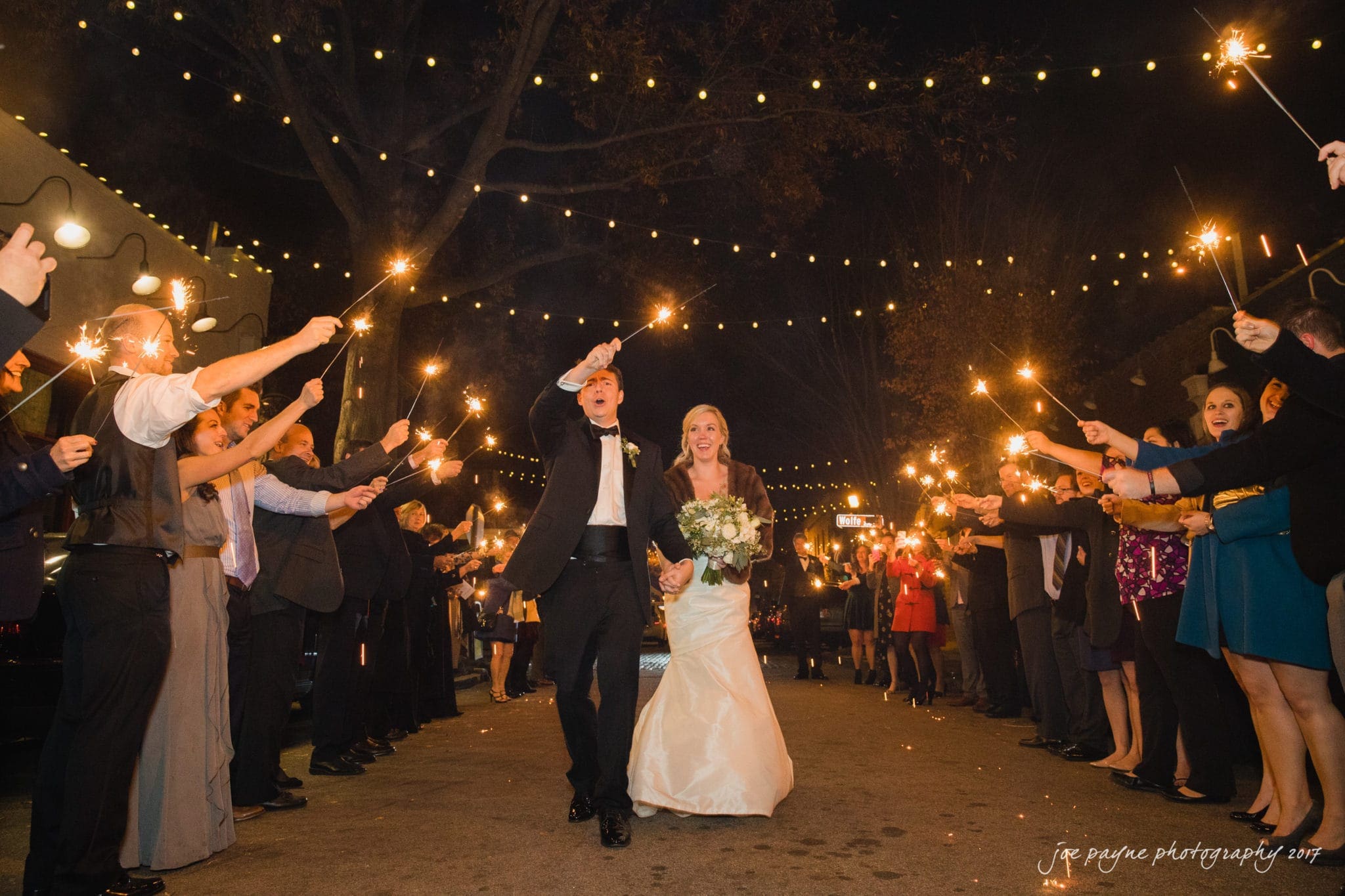 A bride and groom celebrate at night, walking down a street lined with guests holding sparklers. The bride wears a white dress and holds a bouquet, while the groom is in a tuxedo. String lights are visible above, creating a festive atmosphere.