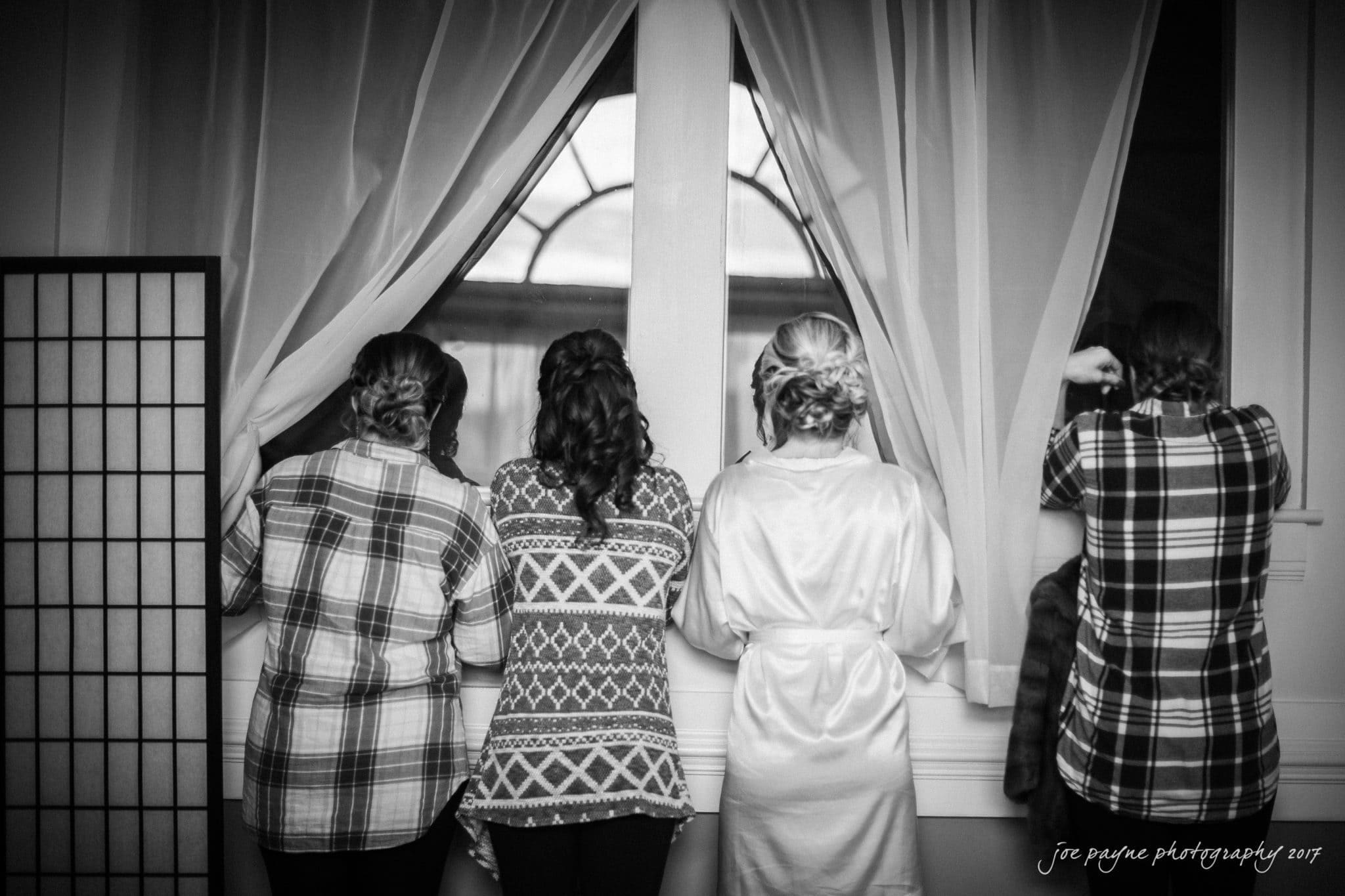 Four women standing and looking out a window with sheer curtains, dressed in casual and patterned attire, including a robe. The setting appears to be indoors with a peaceful, contemplative mood. Black and white photograph.
