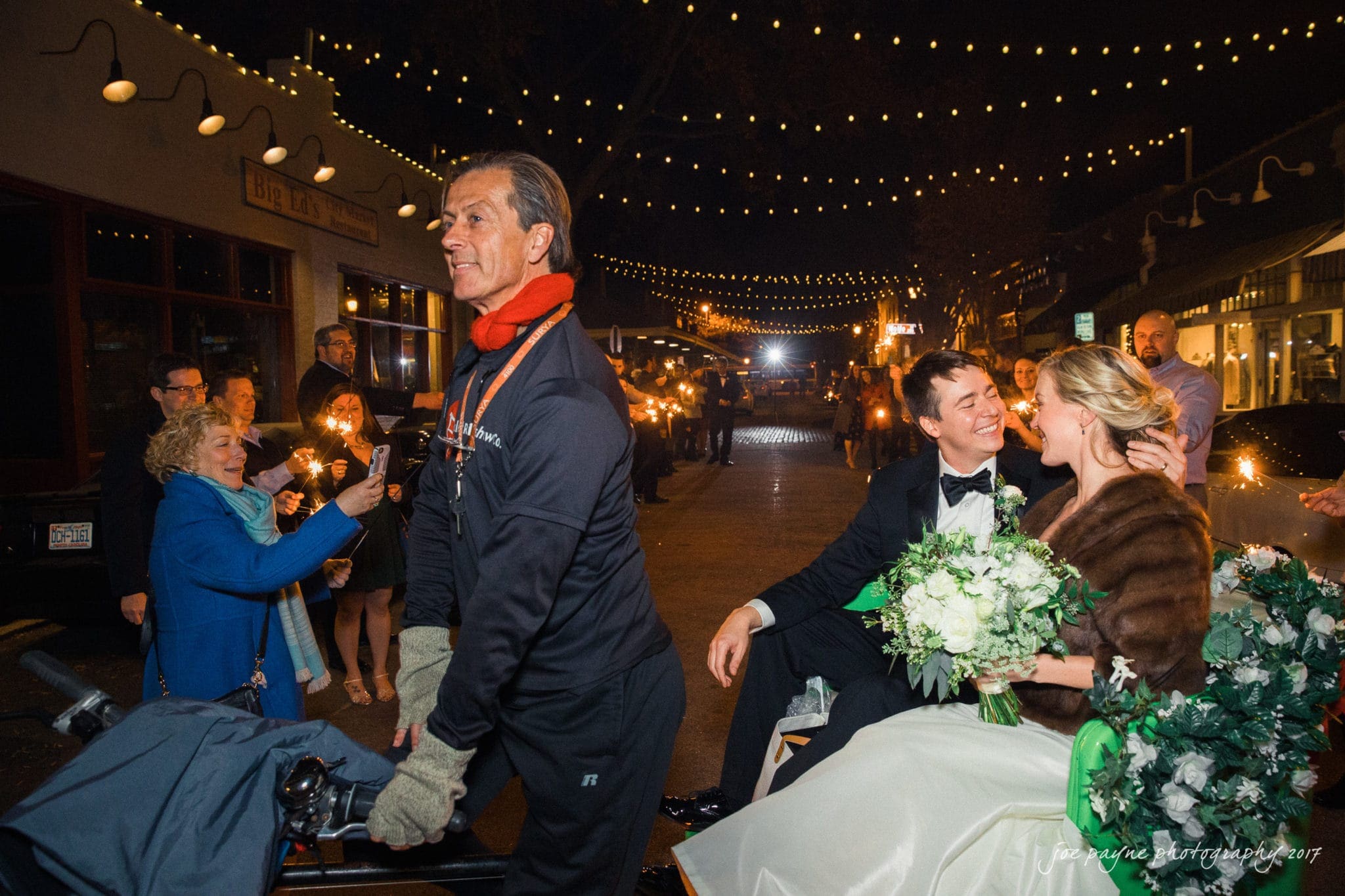 A newlywed couple sits on a rickshaw decorated with flowers at night. The bride holds a bouquet, and the groom wears a tuxedo. Lights hang above, and onlookers with sparklers surround them. A man in a warm outfit pulls the rickshaw through the street.