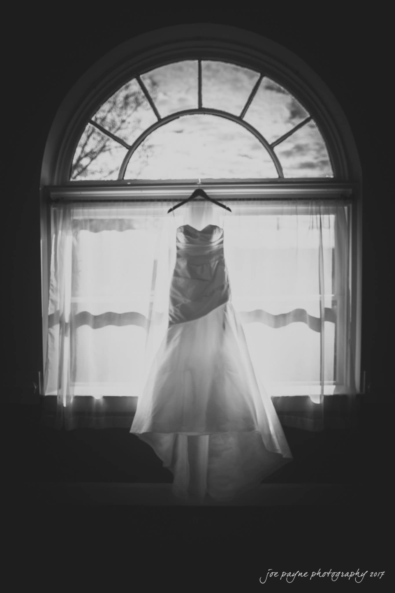 A black and white photo of a wedding dress hanging in front of a large arched window with curtains. The dress is illuminated by the light coming through the window, highlighting its elegant design against the dark background.