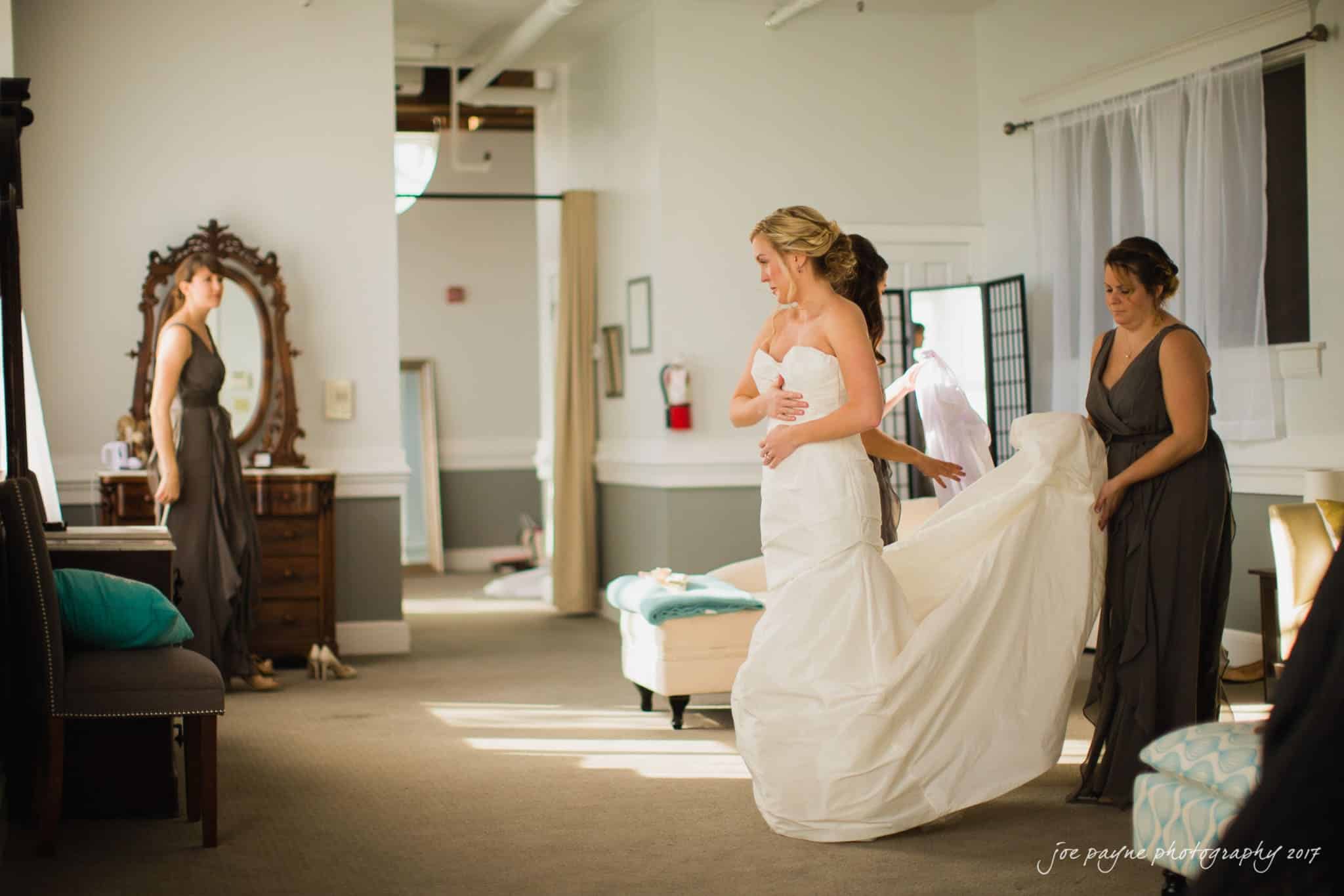 A bride in a white wedding dress is being assisted by two bridesmaids in a warmly lit room. One bridesmaid is adjusting the train of the dress while the other is standing by a vintage dresser. A large mirror and various furniture pieces are visible.