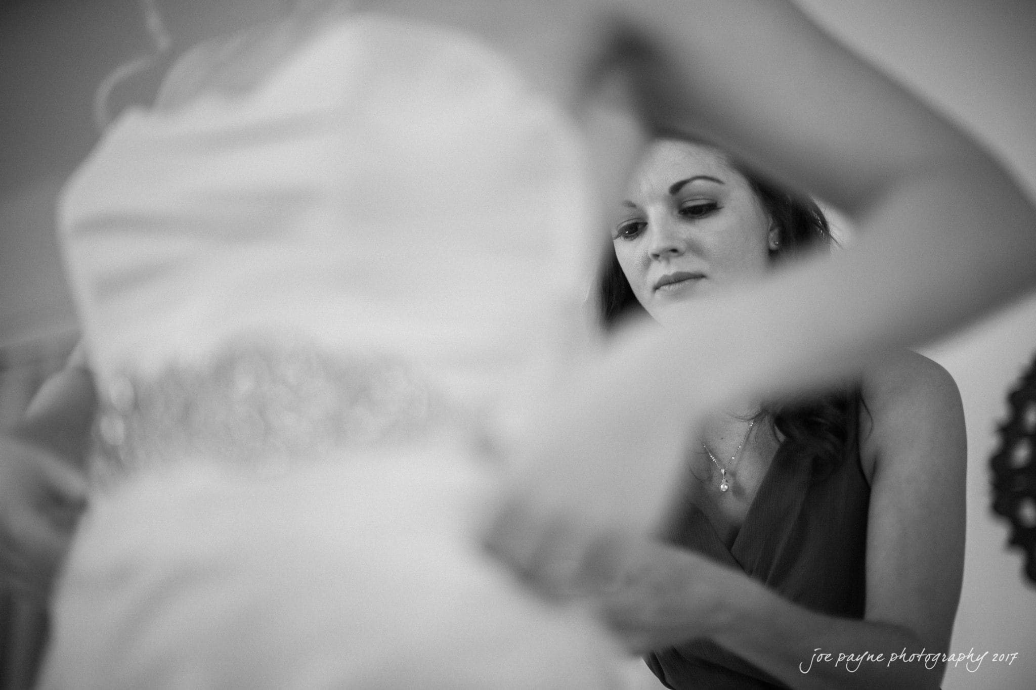 A woman in a dark dress assists someone wearing a white dress, focusing on the intricate details. The image captures a moment of preparation in a softly lit setting.