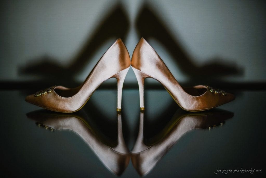 Elegant pink high heels with decorative elements on the toes are positioned back to back, casting symmetrical shadows on a reflective surface against a neutral background.