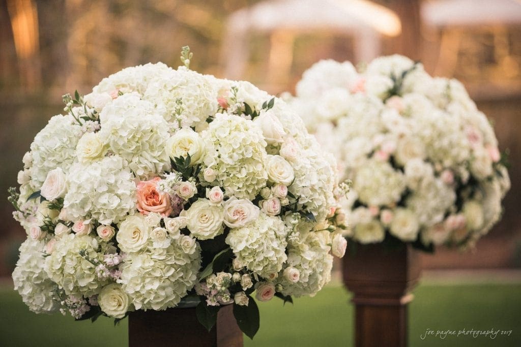 Two large floral arrangements featuring white hydrangeas and soft pink roses are displayed on wooden pedestals outdoors. The background is softly blurred, creating a romantic and elegant setting.