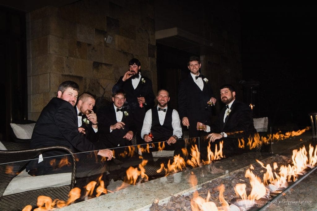 A group of eight men in formal suits are lounging around an outdoor fire pit at night. They are holding drinks and smiling, dressed in tuxedos, suggesting a celebration or wedding event. The fire casts a warm glow on their faces.