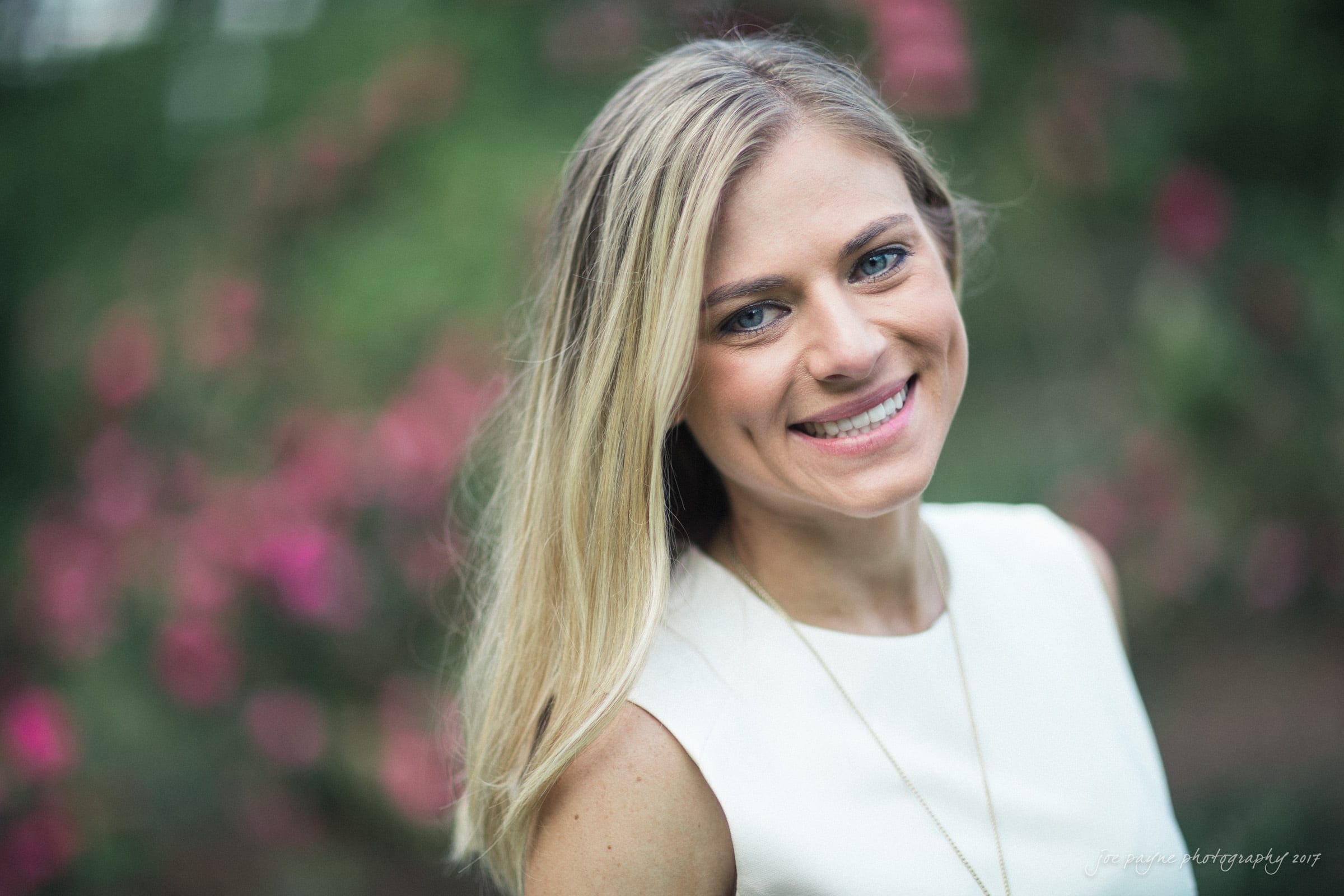 duke gardens engagement photography - margo & patrick A smiling woman with long blonde hair wearing a white sleeveless top stands outdoors, surrounded by blurred pink flowers and greenery in the background.