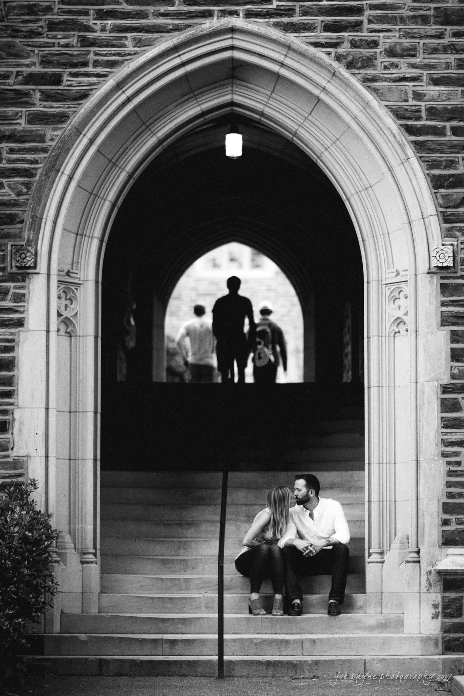 duke gardens engagement photography - margo & patrick A couple sits on the steps of a stone archway, leaning in for a kiss. In the background, silhouettes of people walking through the corridor are visible. The scene is in black and white, conveying a romantic and timeless atmosphere.