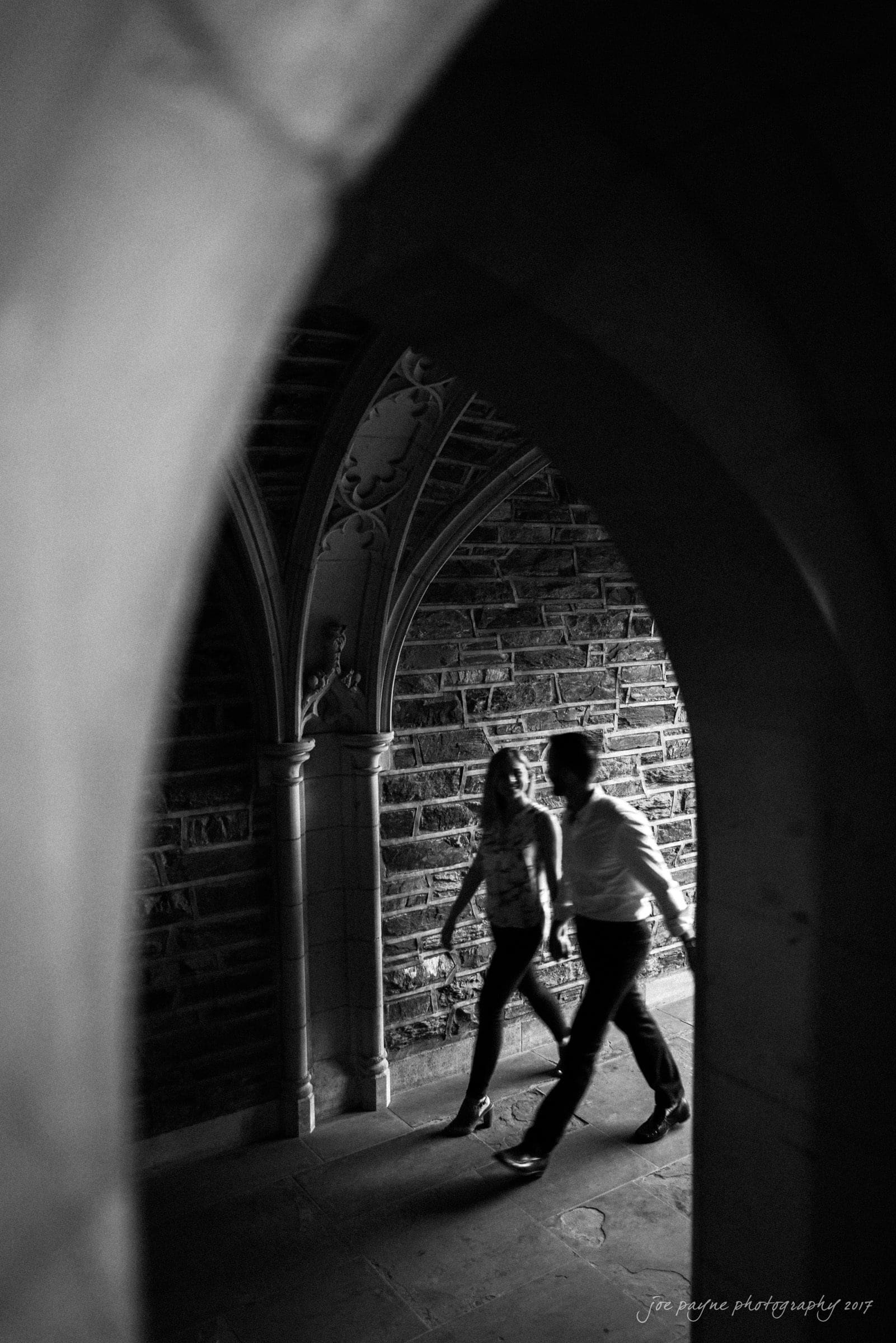 duke gardens engagement photography - margo & patrick A black and white photo of a couple walking through a stone archway in a dimly lit corridor with Gothic architecture. They are slightly blurred, conveying motion. Light and shadows create a dramatic effect.