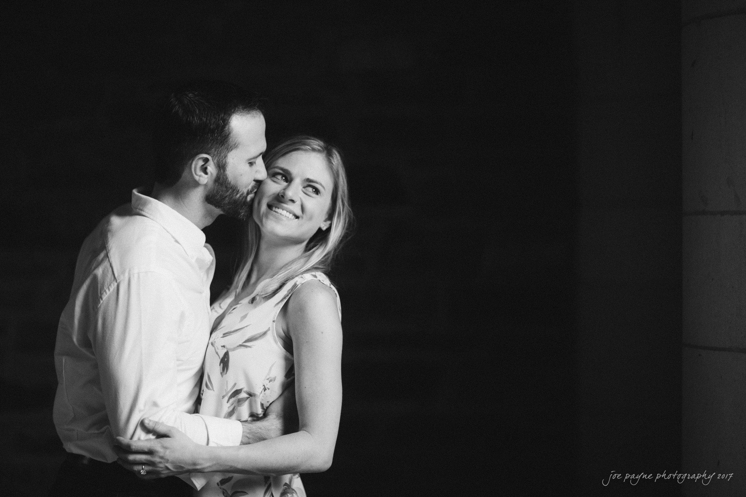 duke gardens engagement photography - margo & patrick A black and white image of a couple embracing. The man, wearing a white shirt, gently kisses the smiling woman on the cheek. She wears a sleeveless floral top, looking upward with a happy expression. The background is dark and blurred.