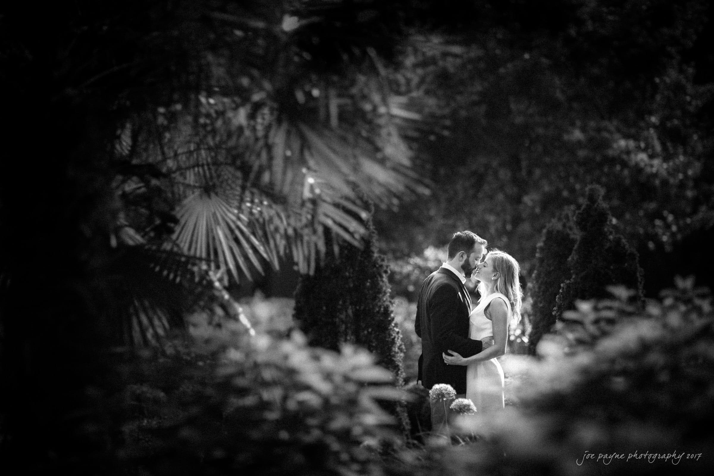 duke gardens engagement photography - margo & patrick A couple stands closely together, gazing into each others eyes in a lush garden setting. Soft dappled sunlight filters through the foliage, creating a romantic, intimate atmosphere. The image is in black and white.