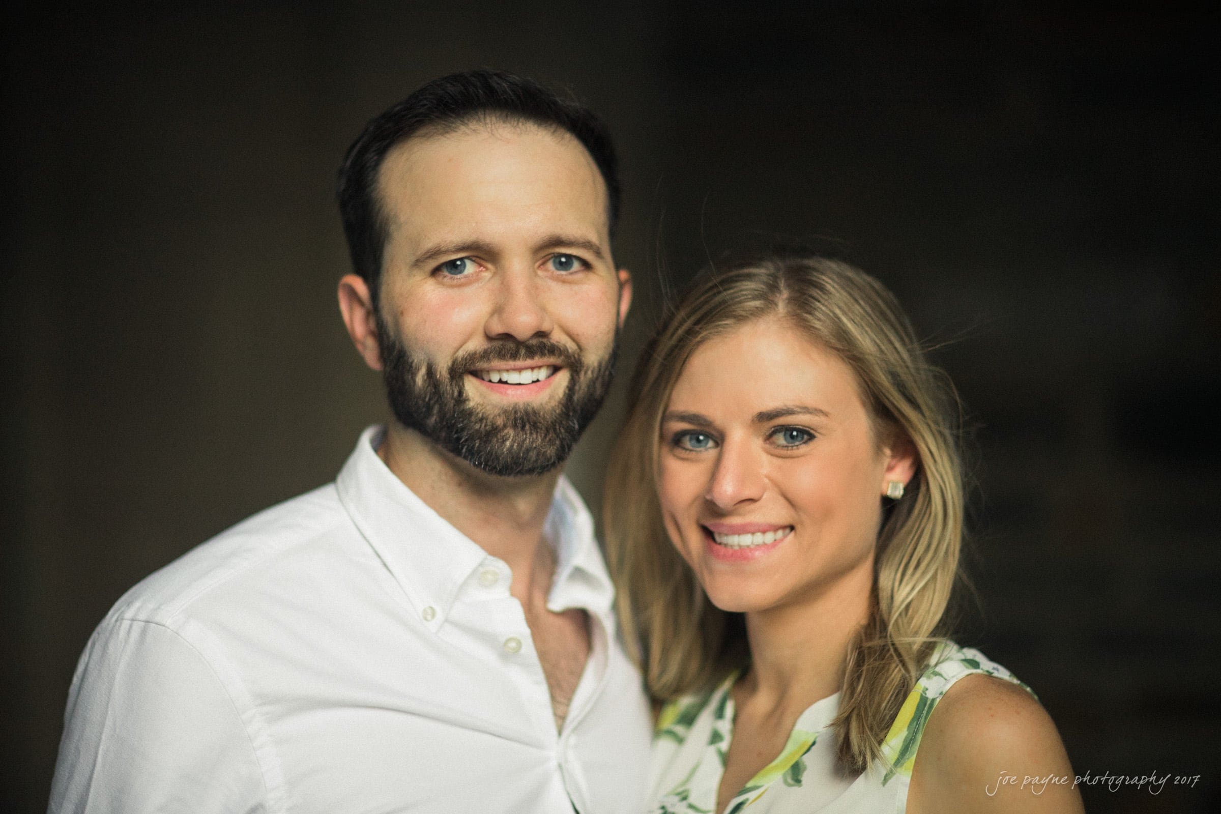 duke gardens engagement photography - margo & patrick A smiling bearded man in a white shirt stands next to a smiling blonde woman in a floral dress, both looking at the camera against a dark, blurred background.