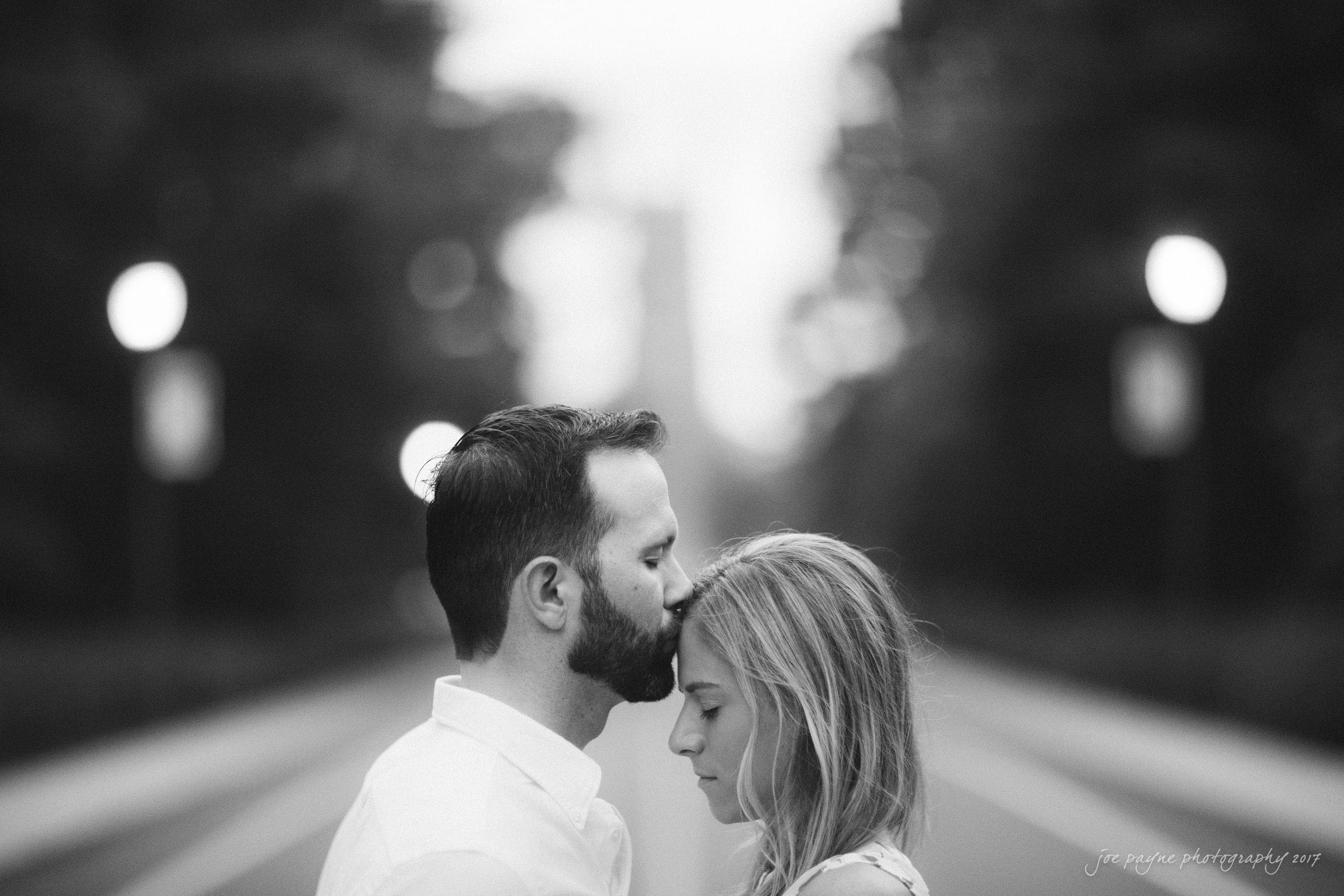 duke gardens engagement photography - margo & patrick A black and white photo of a man gently kissing a womans forehead. They stand close together on an empty street lined with blurred lights. The scene is serene and intimate, with a soft background.