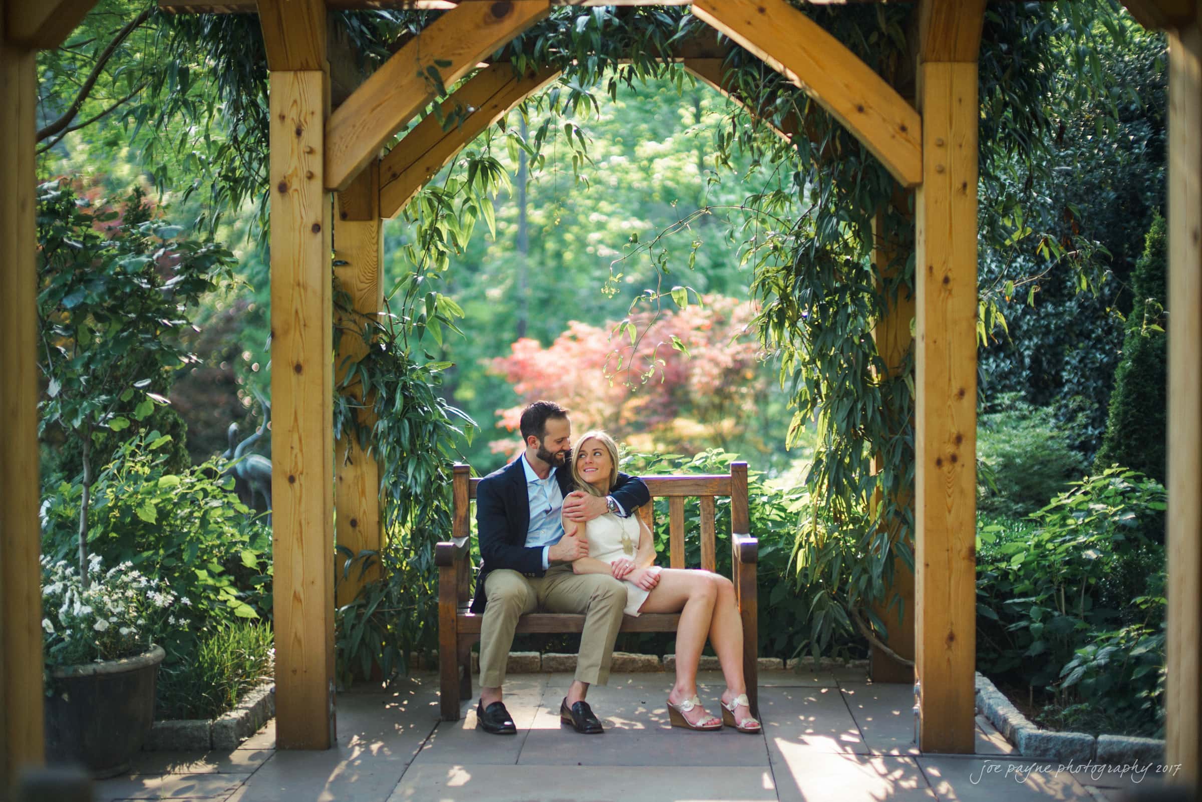 duke gardens engagement photography - margo & patrick A couple sits on a wooden bench under a pergola in a lush garden. They are smiling and cuddling, surrounded by greenery and soft natural light.