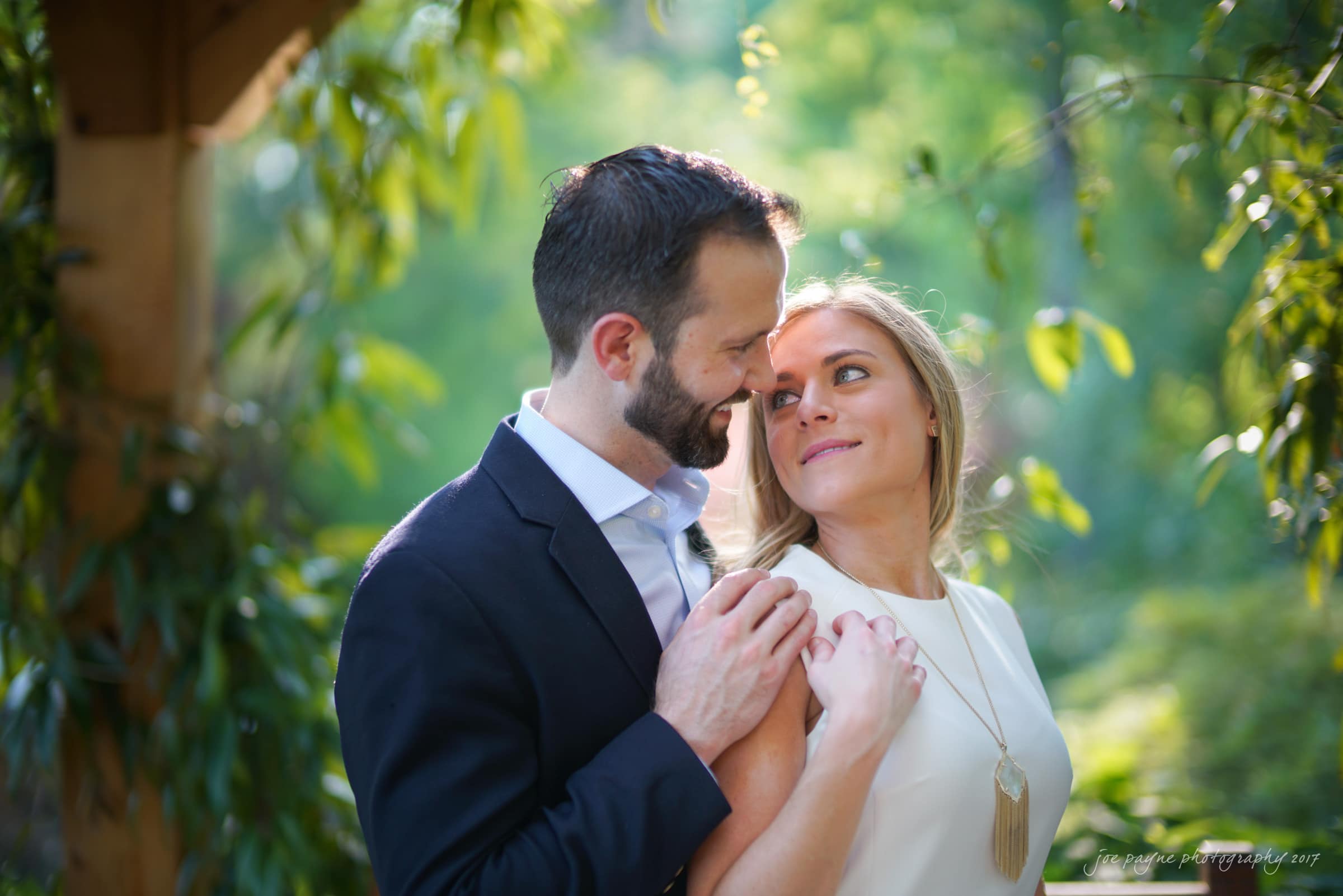 duke gardens engagement photography - margo & patrick A couple stands closely in a lush, green outdoor setting. The man in a dark suit jacket embraces the woman in a white top. They gaze into each others eyes, surrounded by sunlight filtering through the leaves.