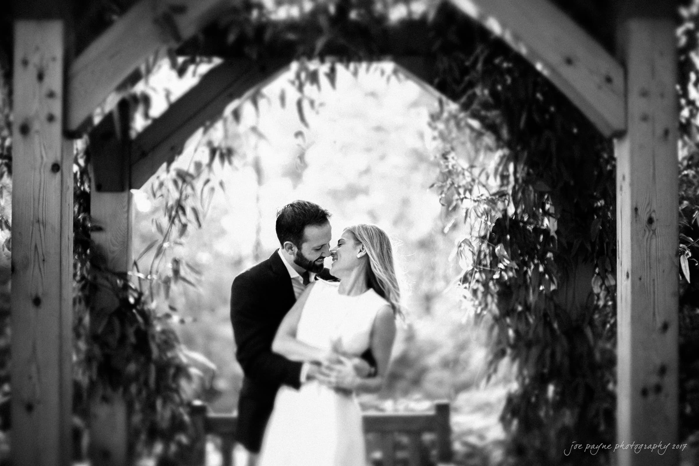 duke gardens engagement photography - margo & patrick A black and white photo of a couple in a loving embrace under a wooden archway surrounded by foliage. They are dressed formally, with the woman in a white dress and the man in a suit, sharing a joyful moment.