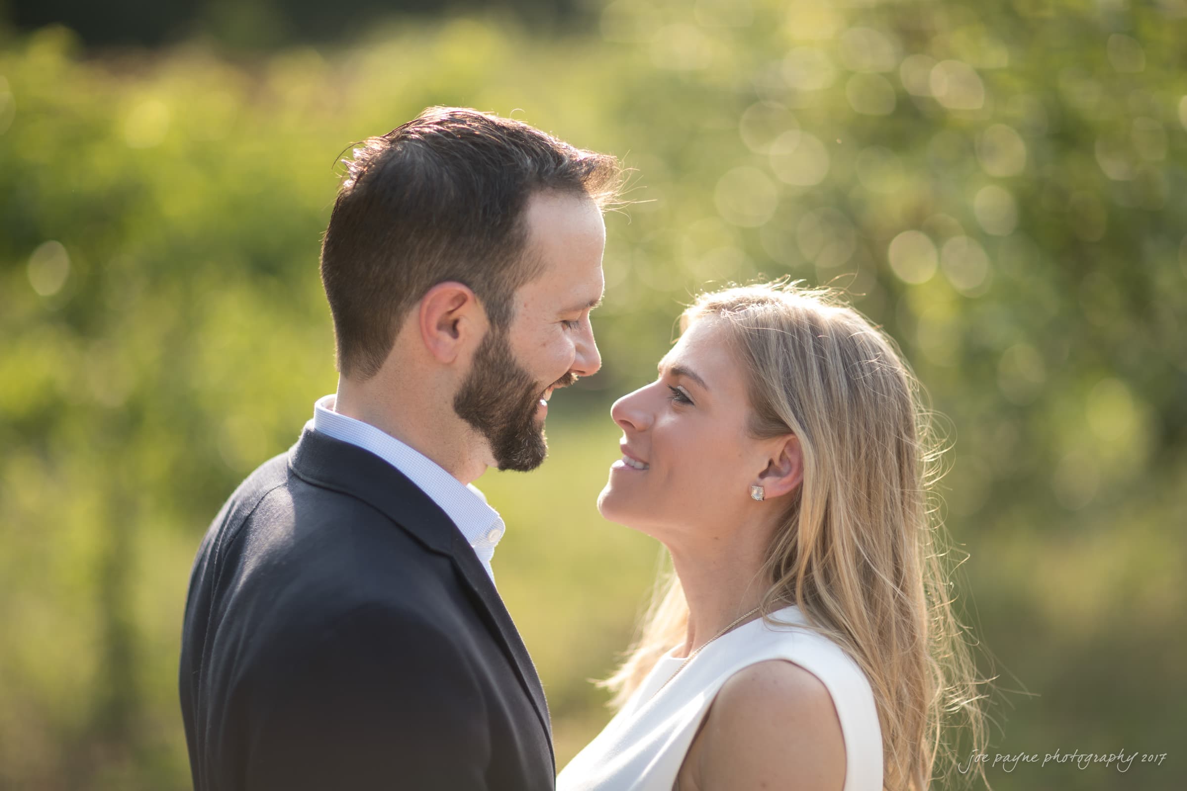 duke gardens engagement photography - margo & patrick A man and woman stand closely, smiling at each other in a sunlit outdoor setting with blurred greenery in the background. The man wears a dark suit, and the woman is in a white sleeveless top.
